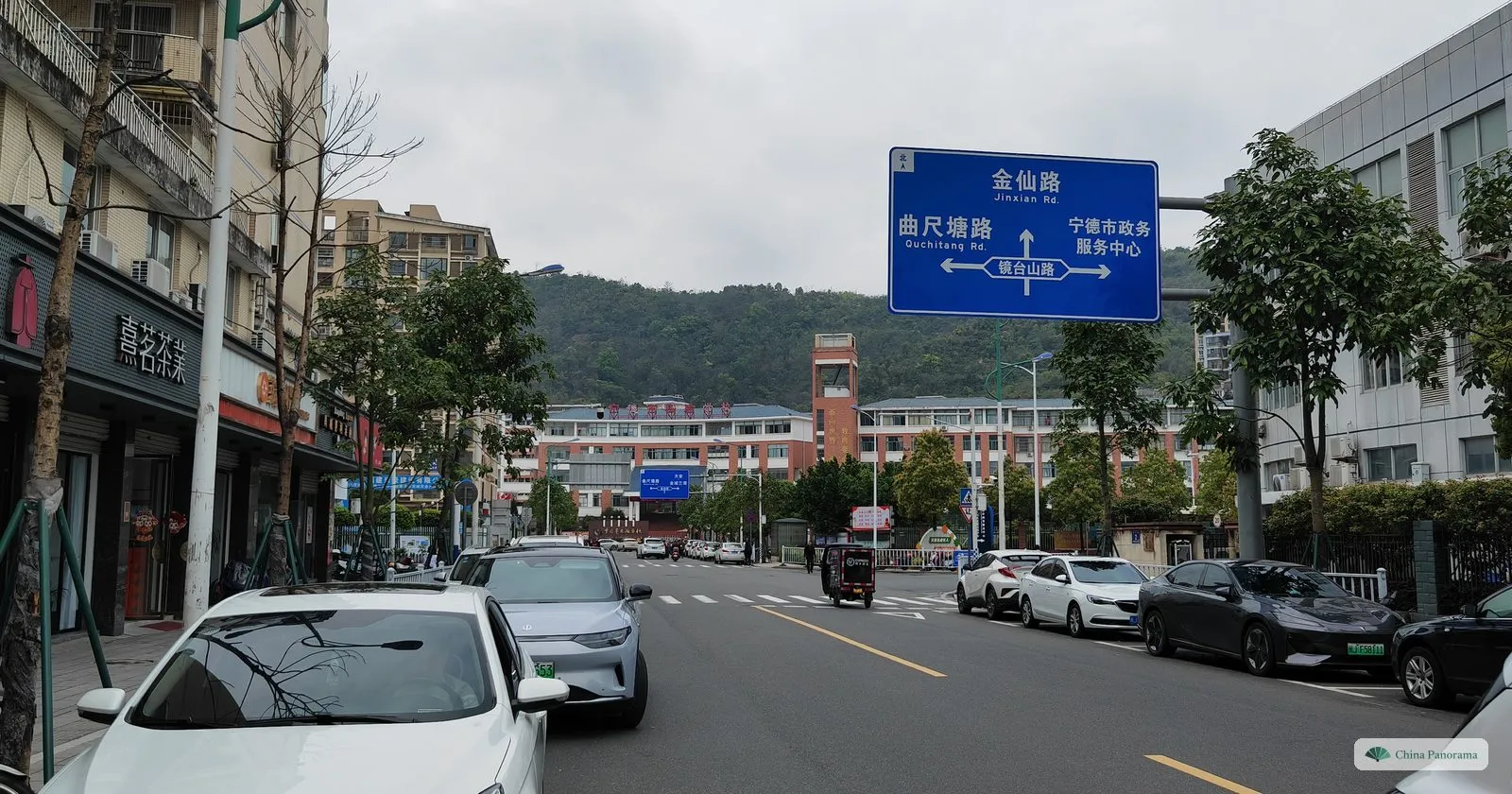 A city street with parked cars, shops on the left and a blue overhead sign in Chinese and English naming Jinxian Road and Quchitang Road and pointing to the city’s government service centre, a school and a wooded hill behind