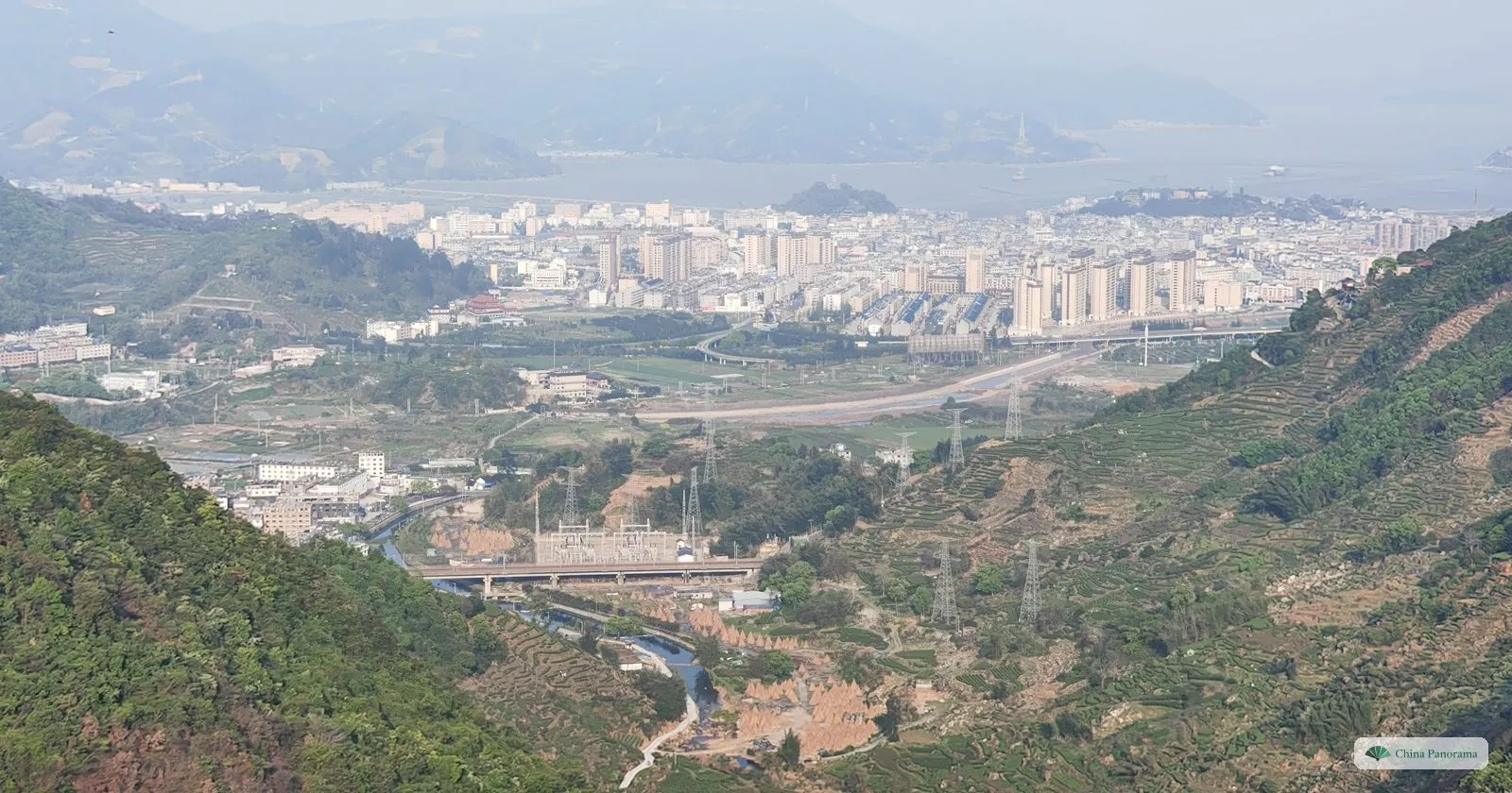 A view down a valley of terraced tea to a town of tower blocks on a coastal plain, a grey bay with small islands beyond and hazy mountains on the far shore