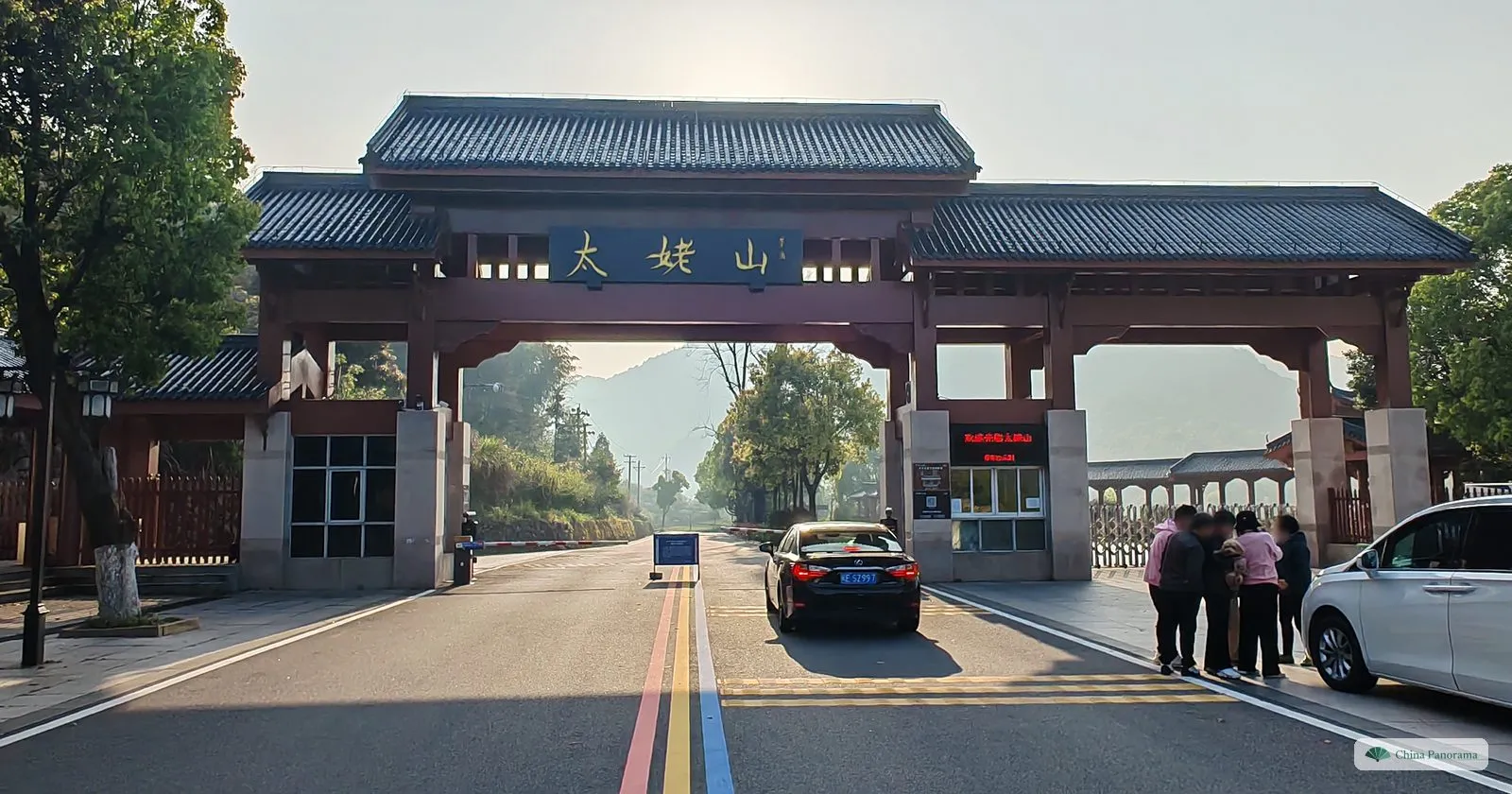 A wide timber gateway with grey tiled roofs and a dark board reading 太姥山 over a road painted with red, yellow and blue lines, a car at the ticket booth and a group of people beside a white van
