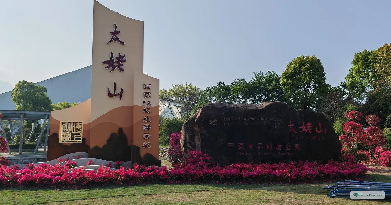 A tall beige sign with the red characters 太姥山 and a vertical line reading 国家5A级旅游景区, a gold seal, and beside it a large dark boulder inscribed 宁德世界地质公园, all behind a bed of magenta bougainvillea