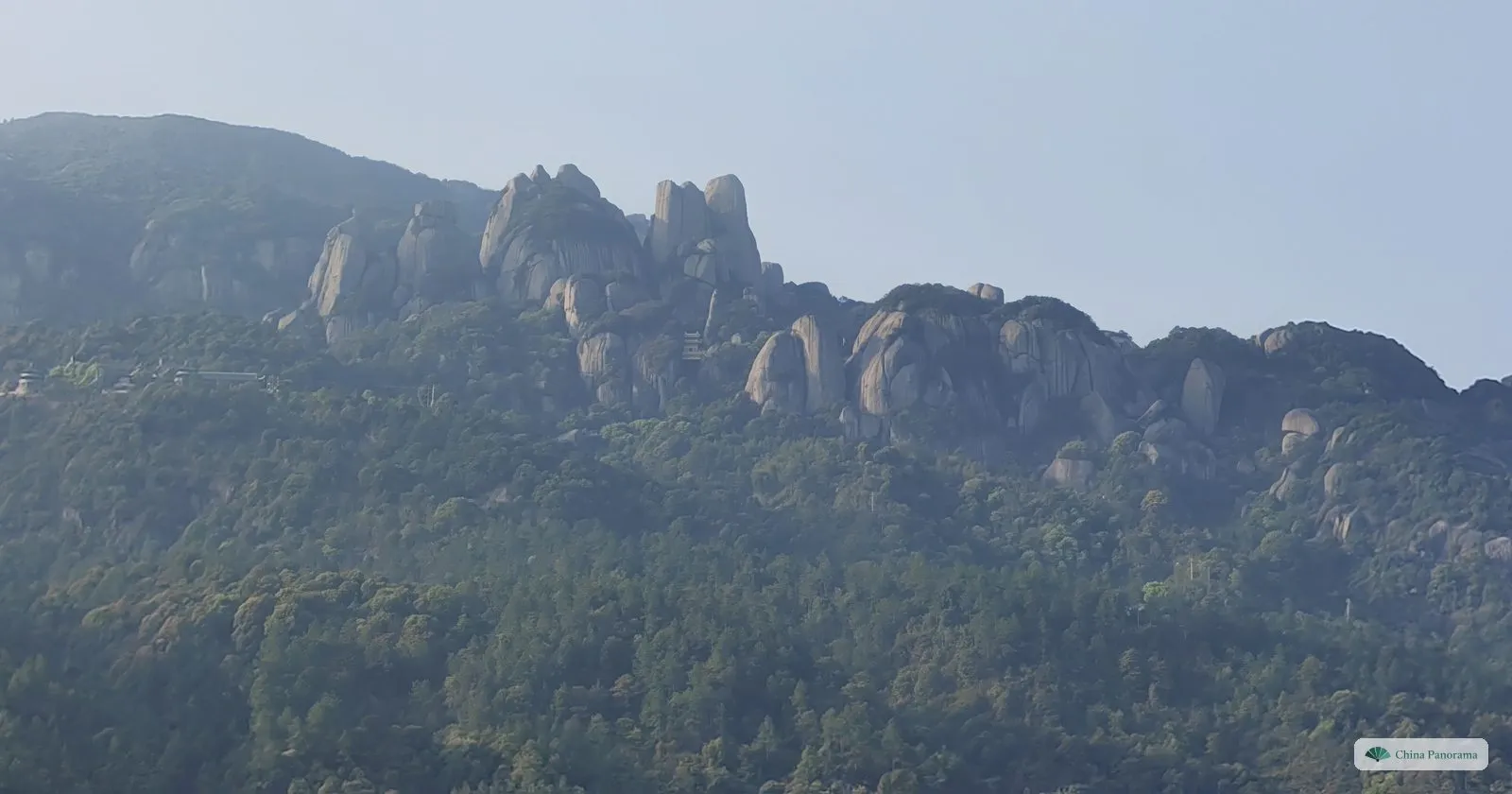 A forested mountainside with a ridge of tall rounded granite towers in spring haze