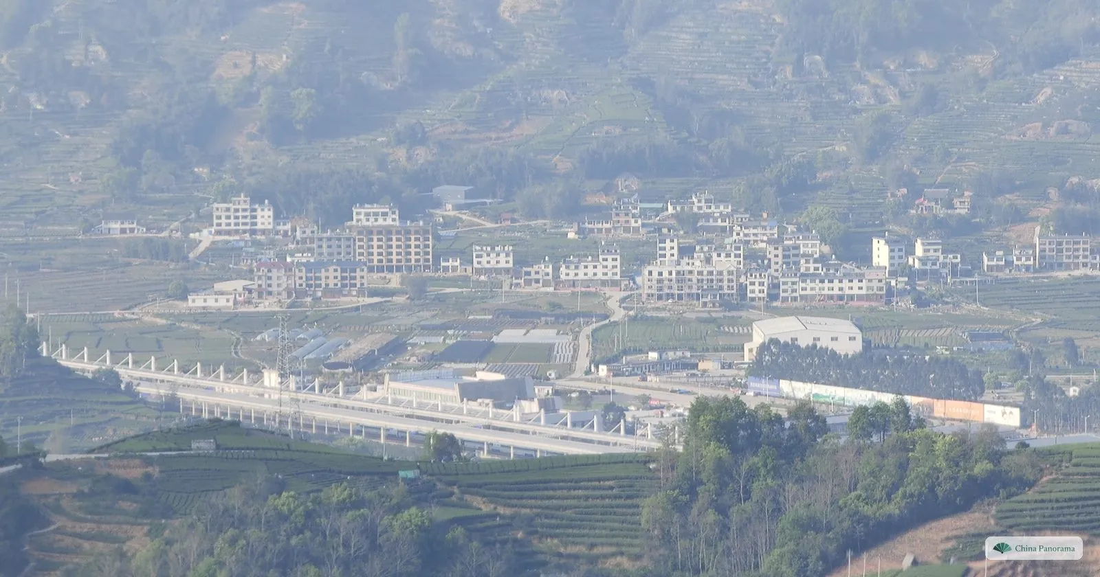 An elevated railway station among tea terraces with a village of white houses behind