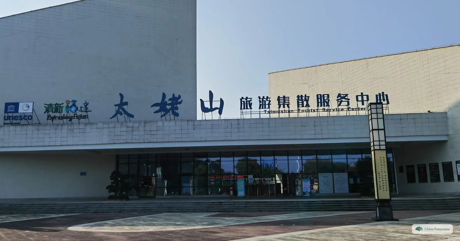 A white building with large blue characters reading 太姥山旅游集散服务中心 and an English line Taimushan Tourist Service Center, a UNESCO geopark logo and a glass entrance above a wide paved forecourt