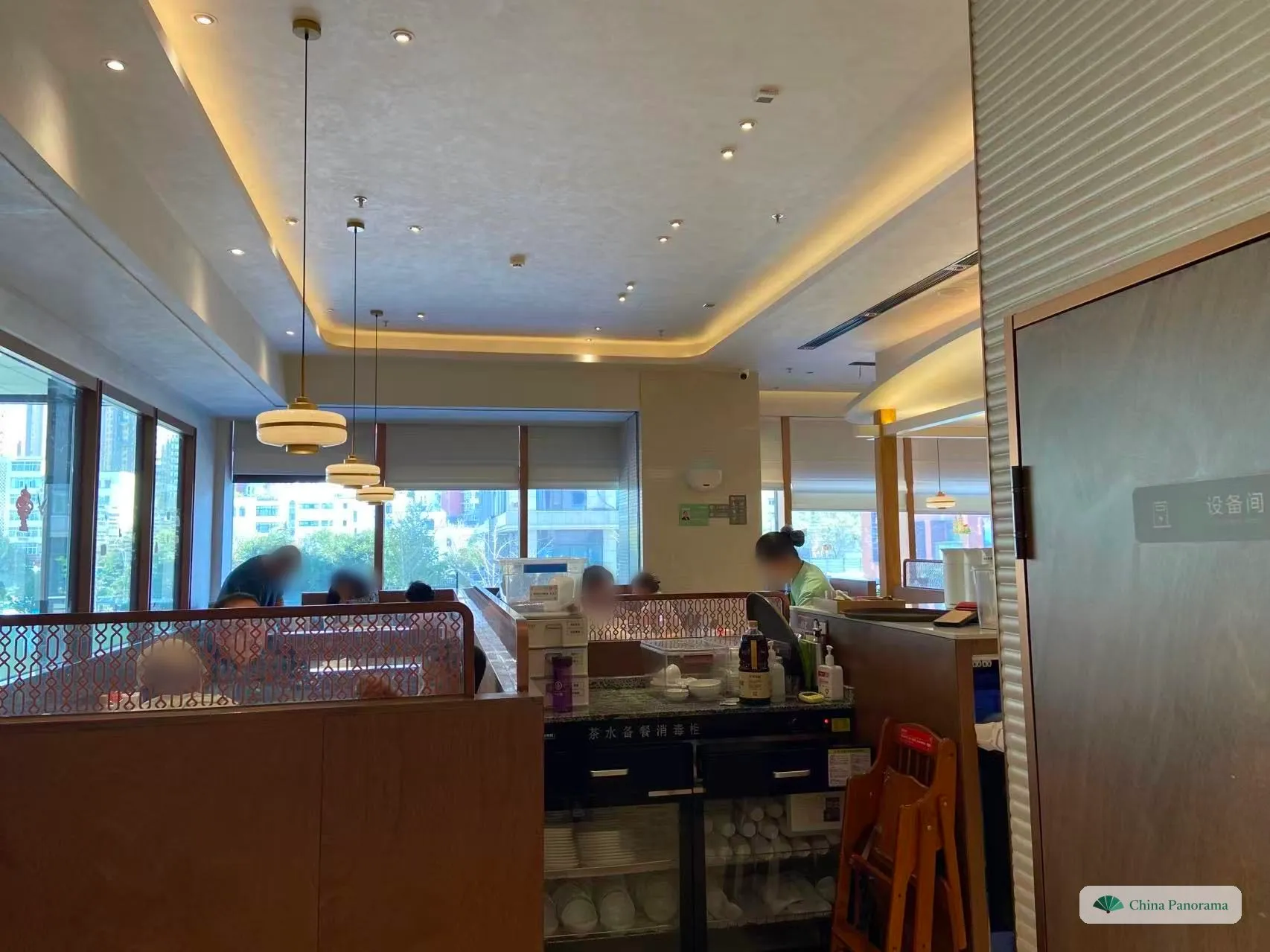 A restaurant dining room with booths behind lattice screens, large windows, a waitress at a service station and a sterilising cabinet full of bowls