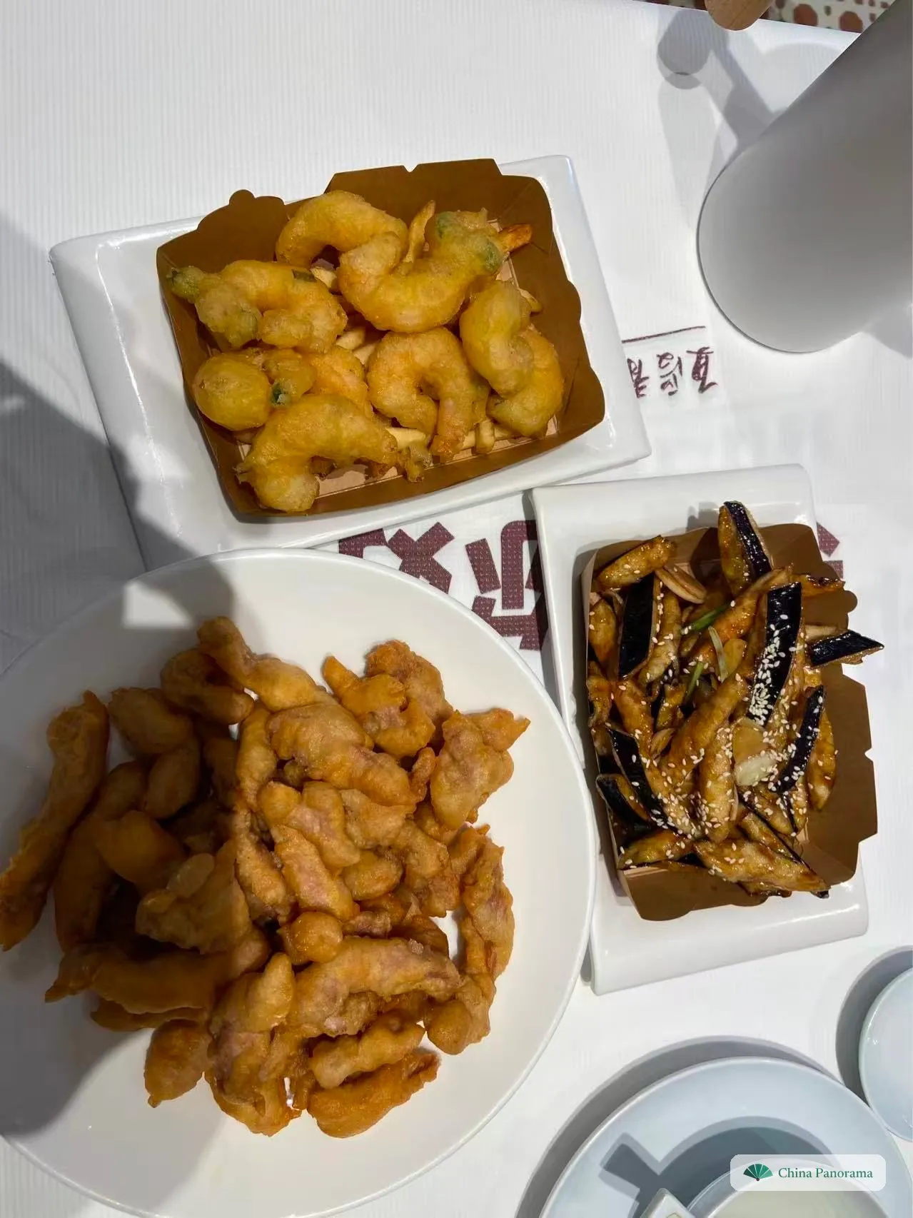 Three dishes seen from above: crispy fried shrimp in a paper-lined tray, aubergine strips with sesame, and a large plate of fried pork tenderloin