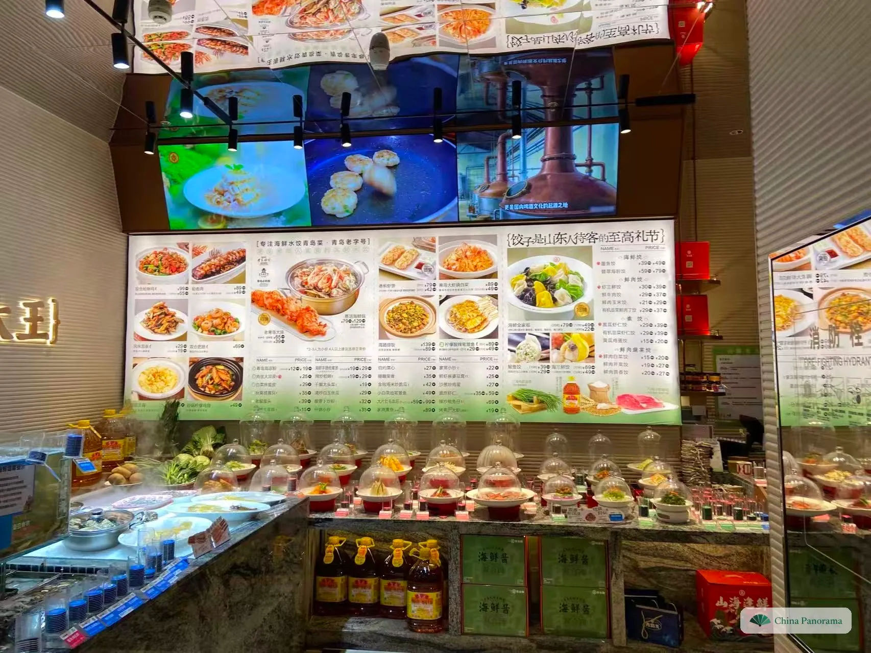The ordering area of a restaurant: a lit picture board with dish photos and prices above two rows of sample dishes under glass domes, raw vegetables on ice on the left