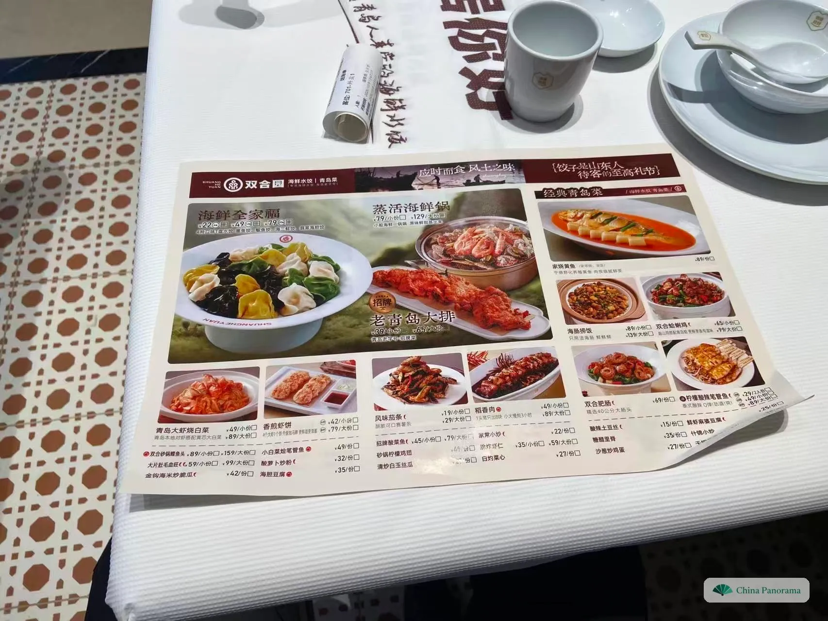 A paper menu on a white tablecloth, with dish photos and a small checkbox next to every price; a tea cup, a bowl with a spoon and a rolled-up printed slip behind it