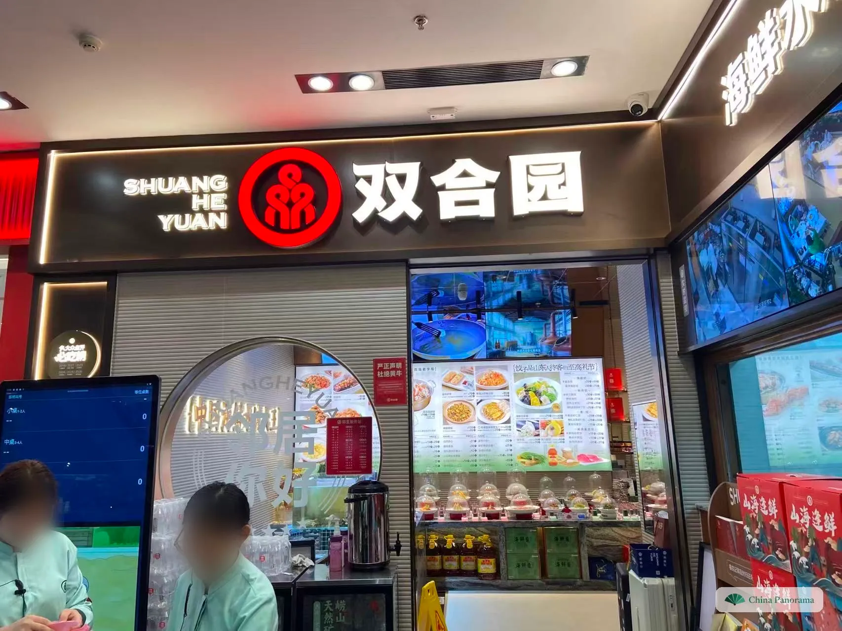 A restaurant entrance in a shopping mall under a lit sign reading 双合园 and SHUANG HE YUAN; inside, a picture menu board above sample dishes under glass domes, and a waiting-list screen on the left