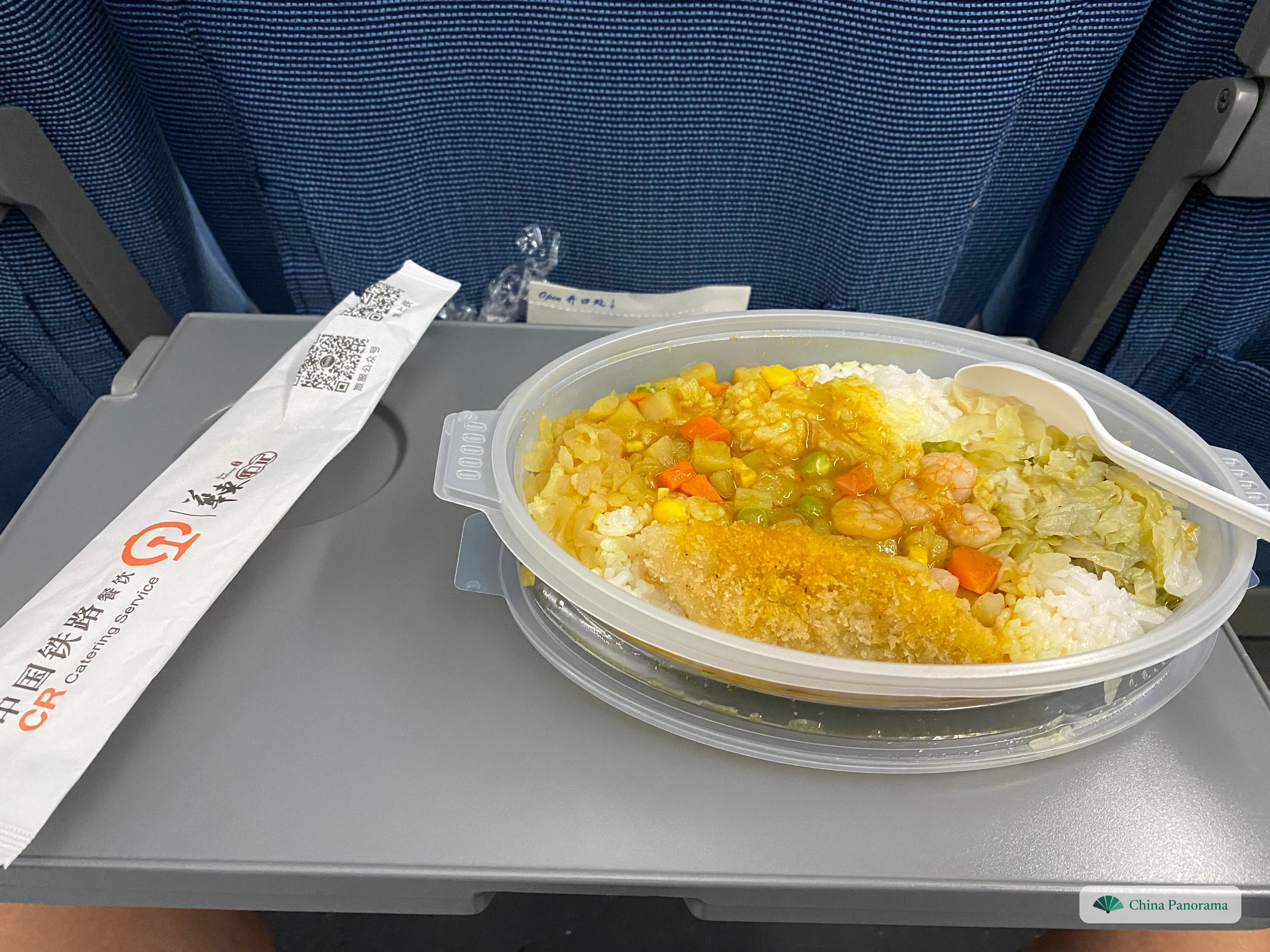 A boxed meal on the fold-down table of a high-speed train: breaded chicken and shrimp curry on rice, next to a China Railway catering chopstick sleeve