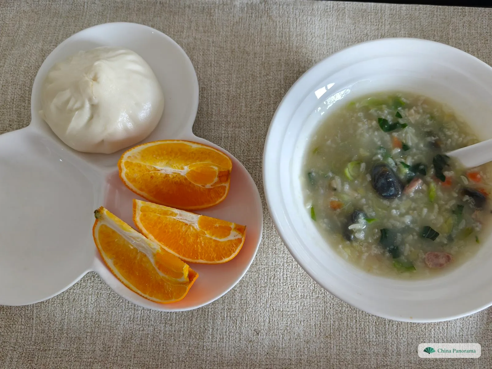 A breakfast from above: a white bowl of congee with century egg, greens, carrot and pork, and next to it a plate with a steamed bun and orange slices