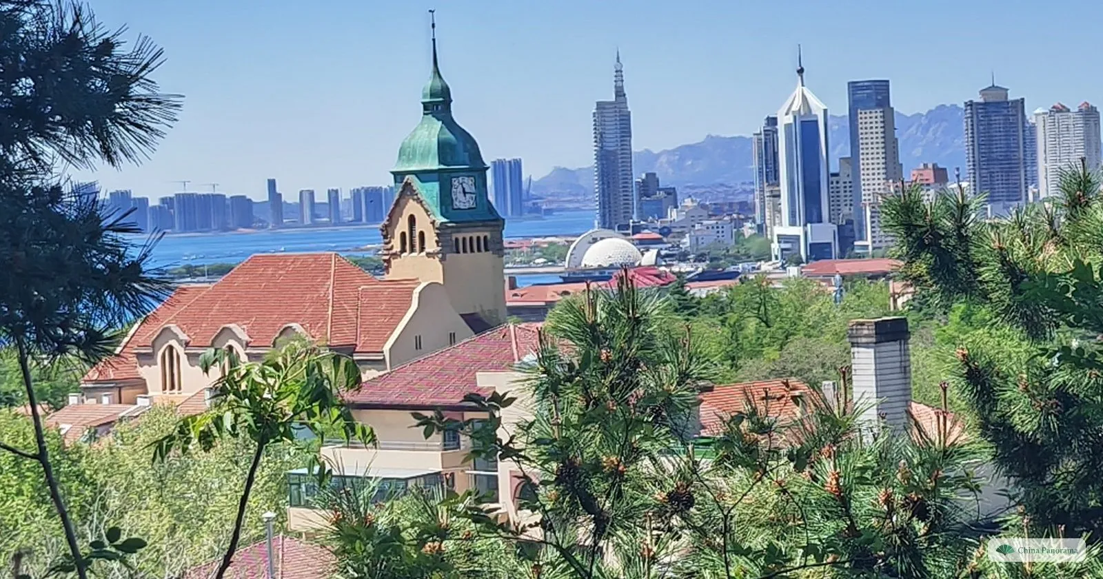 The green clock tower of Qingdao Protestant Church among red roofs, with the modern skyline and the sea behind, seen from Signal Hill