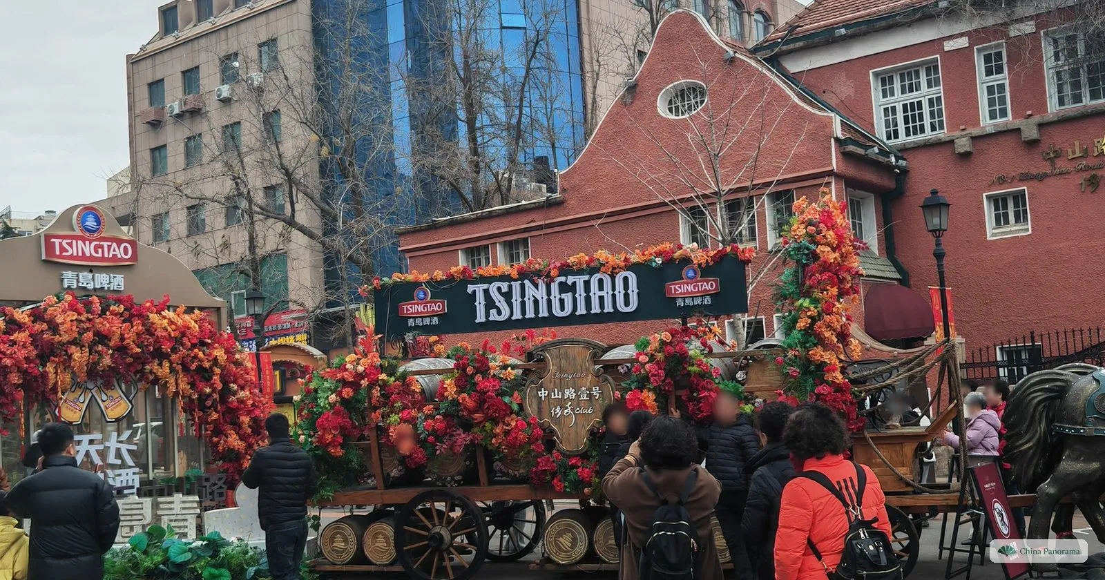 A flower-covered wooden beer wagon under a TSINGTAO sign in front of the red gabled house at No. 1 Zhongshan Road in Qingdao, with visitors taking photos