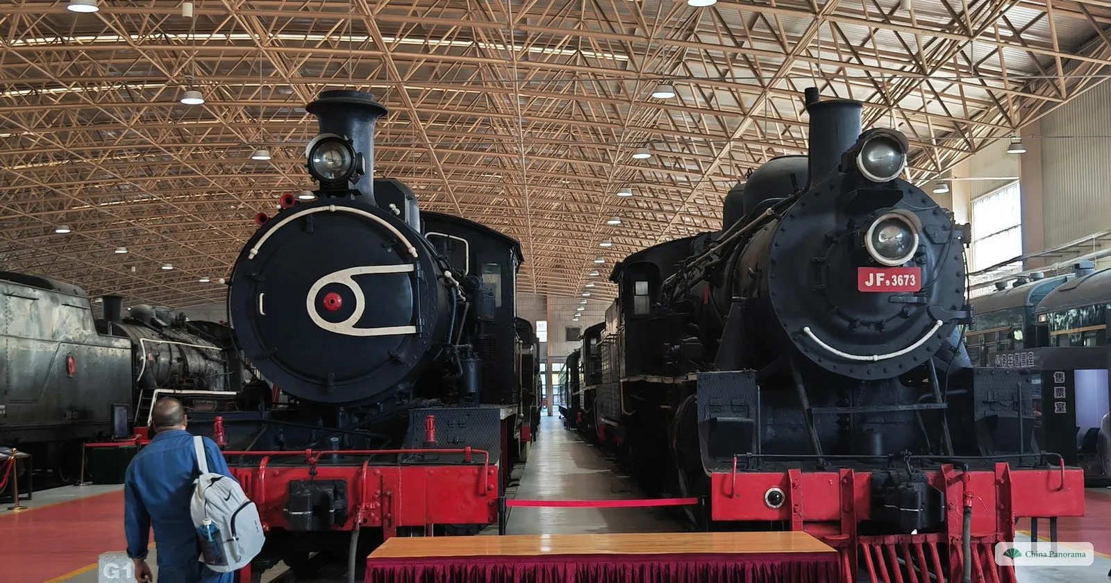 Two early steam locomotives seen head-on on parallel tracks in the China Railway Museum, with a visitor walking between the rails