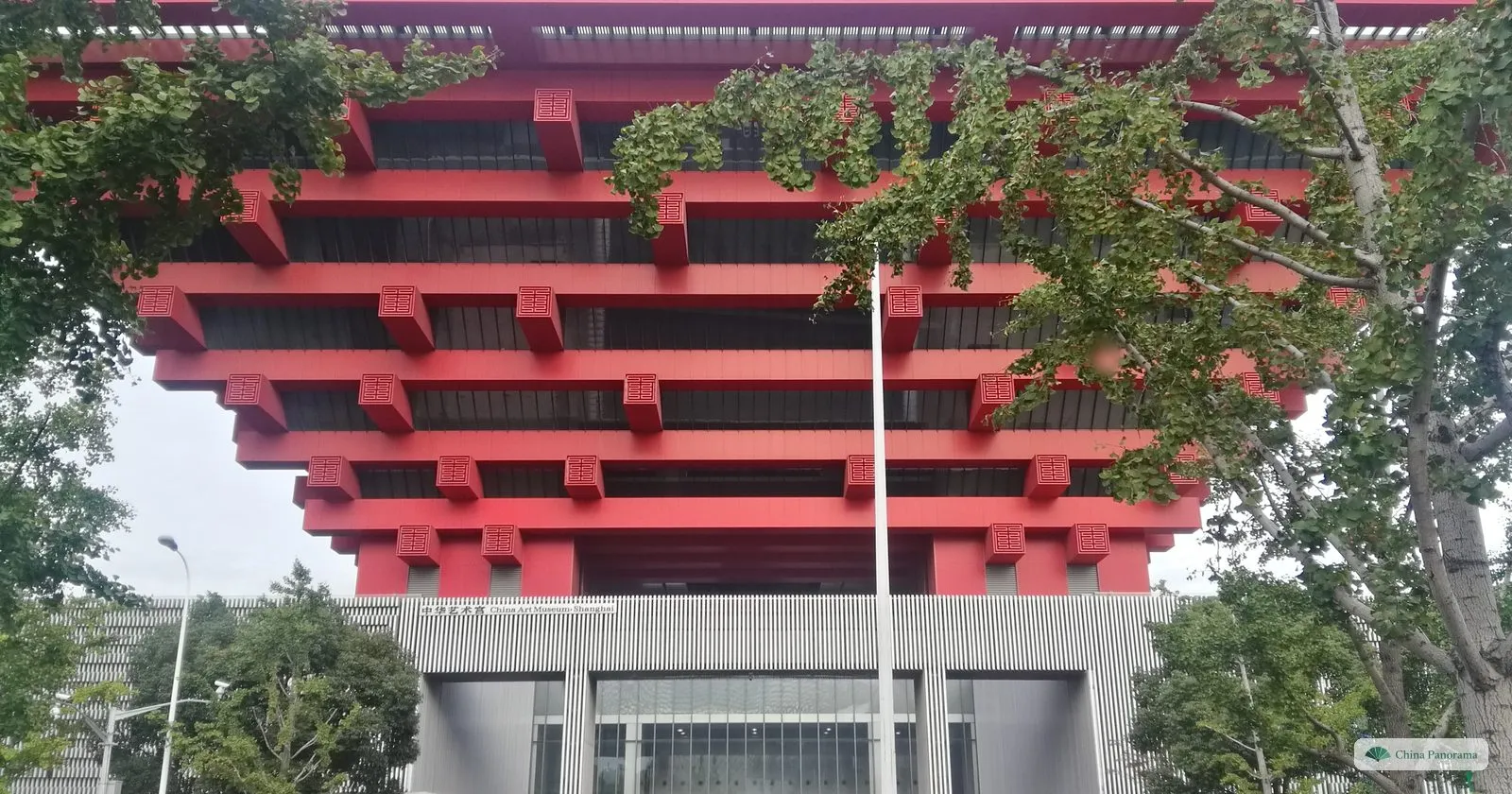 The red stacked beams of the former China Pavilion seen from below, framed by the branches of street trees