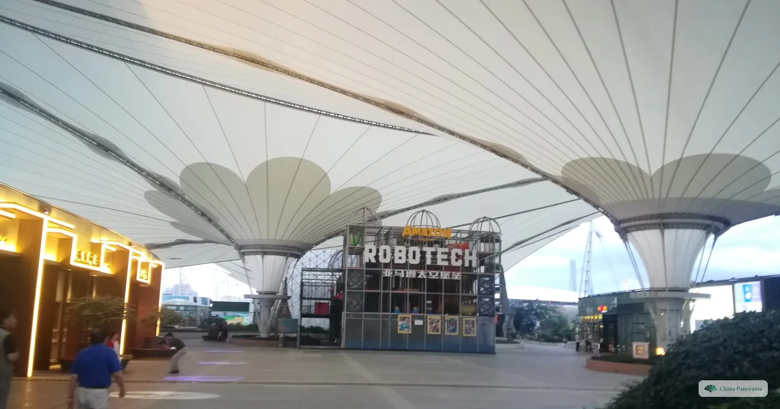 Under the white membrane roof of the Expo Axis at dusk, with two funnel-shaped supports, lit shop fronts and a pavilion reading ROBOTECH