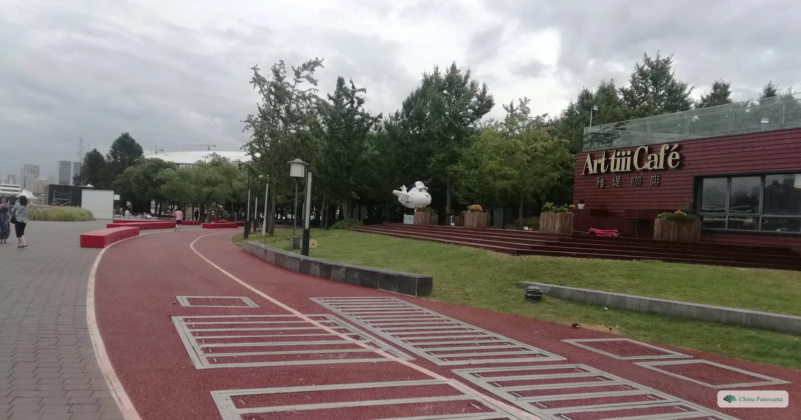 A red running and cycling track through the riverside park on the Expo site, with lawns, trees and a café sign reading Art fiii Café