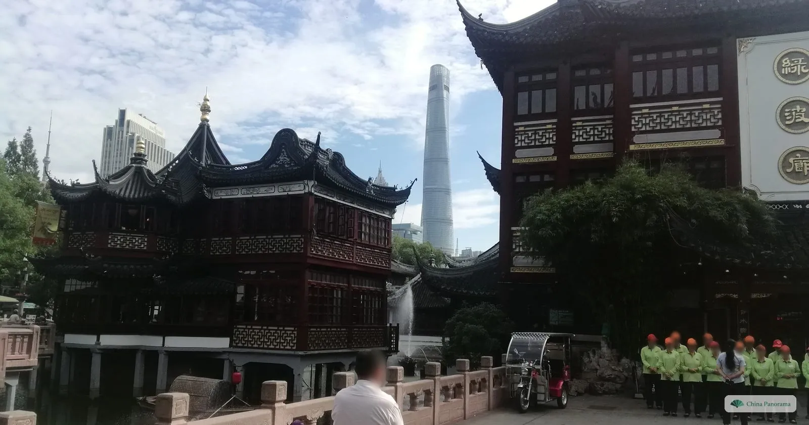 The Huxinting teahouse on stilts in its pond, with its curved black roofs in front of the tall, tapering Shanghai Tower, and staff lined up on the right