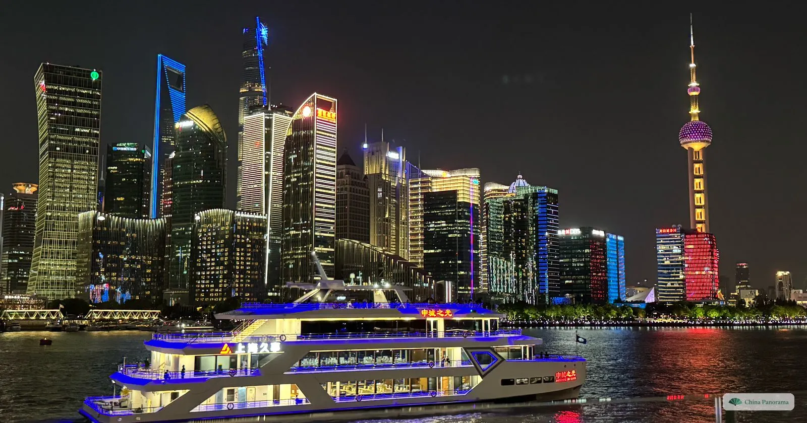 The Lujiazui skyline at night across the Huangpu, with a brightly lit river-cruise boat in front and the Oriental Pearl Tower on the right