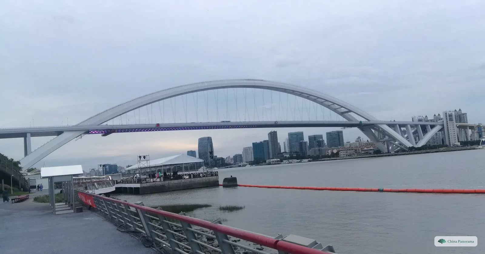 The white steel arch of Lupu Bridge over the Huangpu, seen from the riverside promenade with a red railing