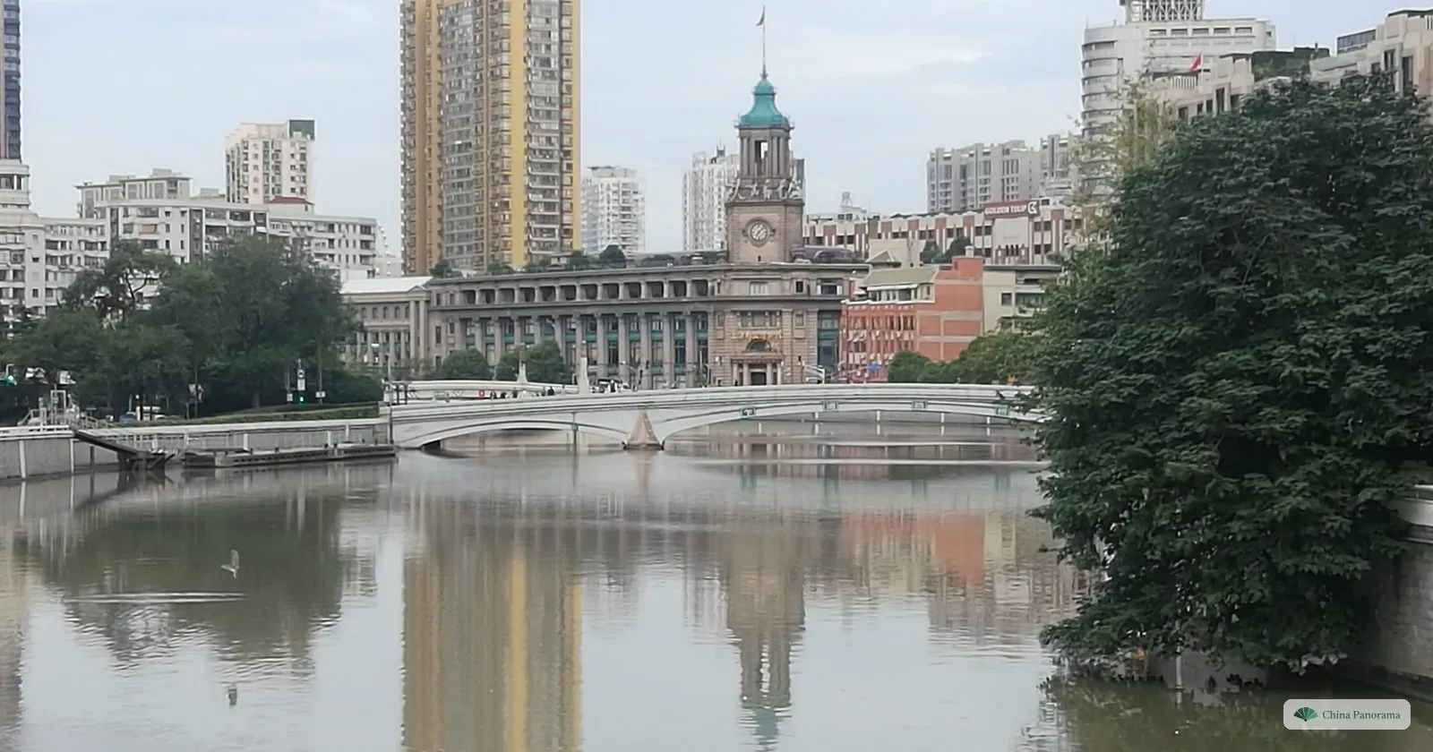The General Post Office on Suzhou Creek, with a clock tower and green cupola, behind a white arched bridge and reflected in the water