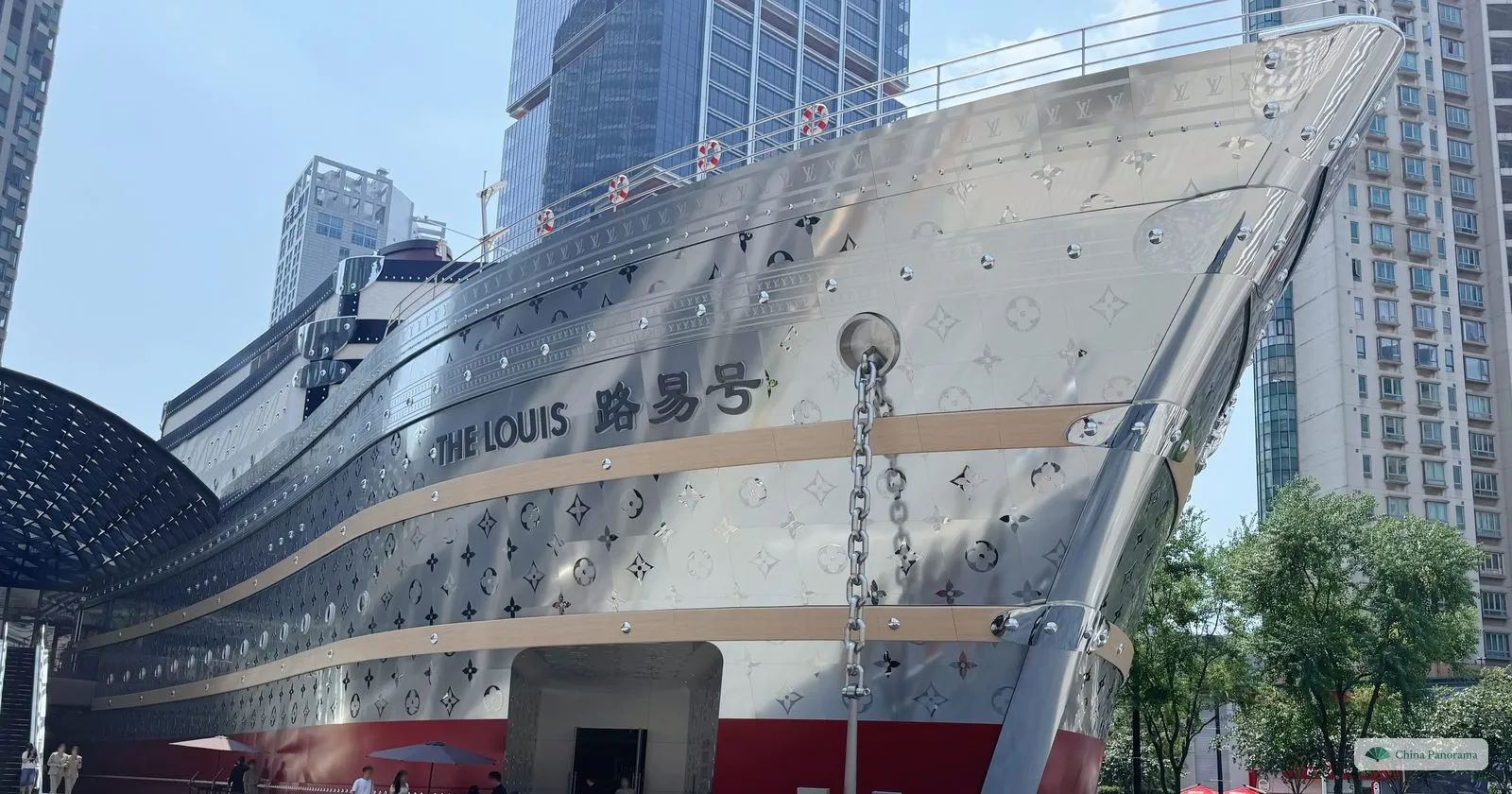 The Louis, a huge steel ship-shaped building with a Monogram pattern, a red hull and an anchor chain, on a plaza between office towers