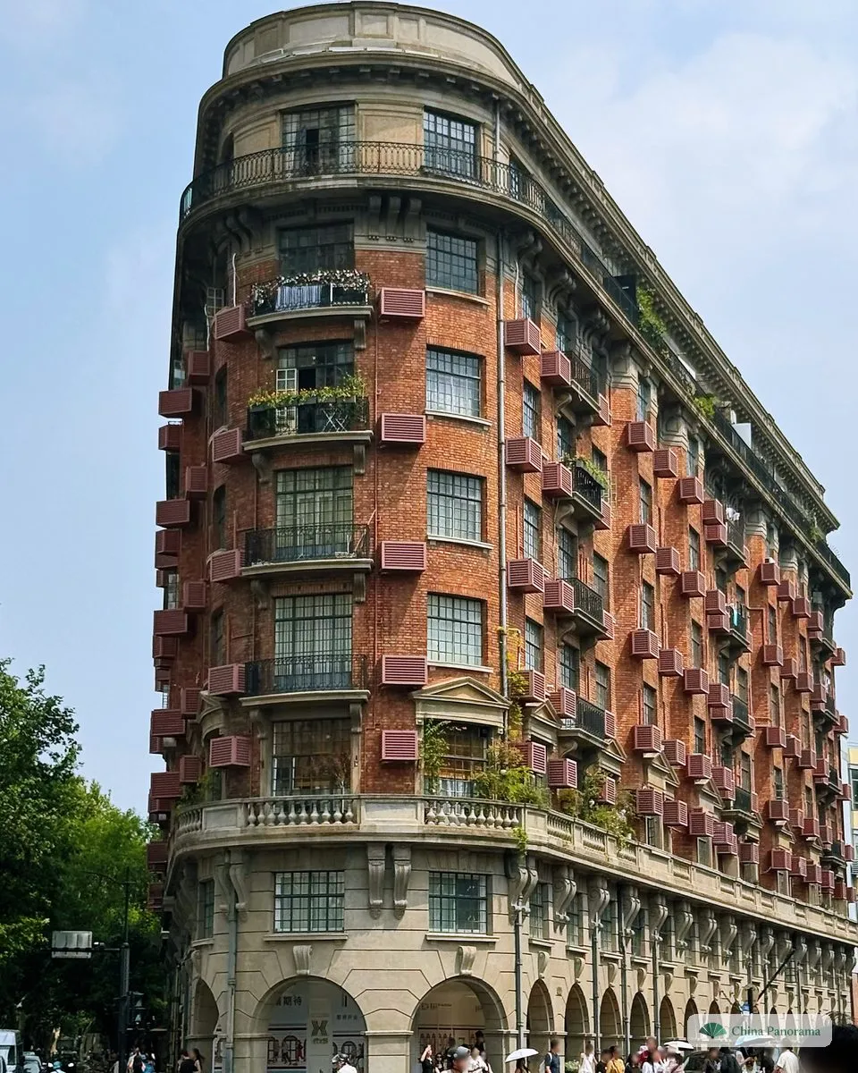 The red-brick Wukang Mansion, a narrow ship-prow-shaped apartment block with balconies, on a street corner with trees and pedestrians