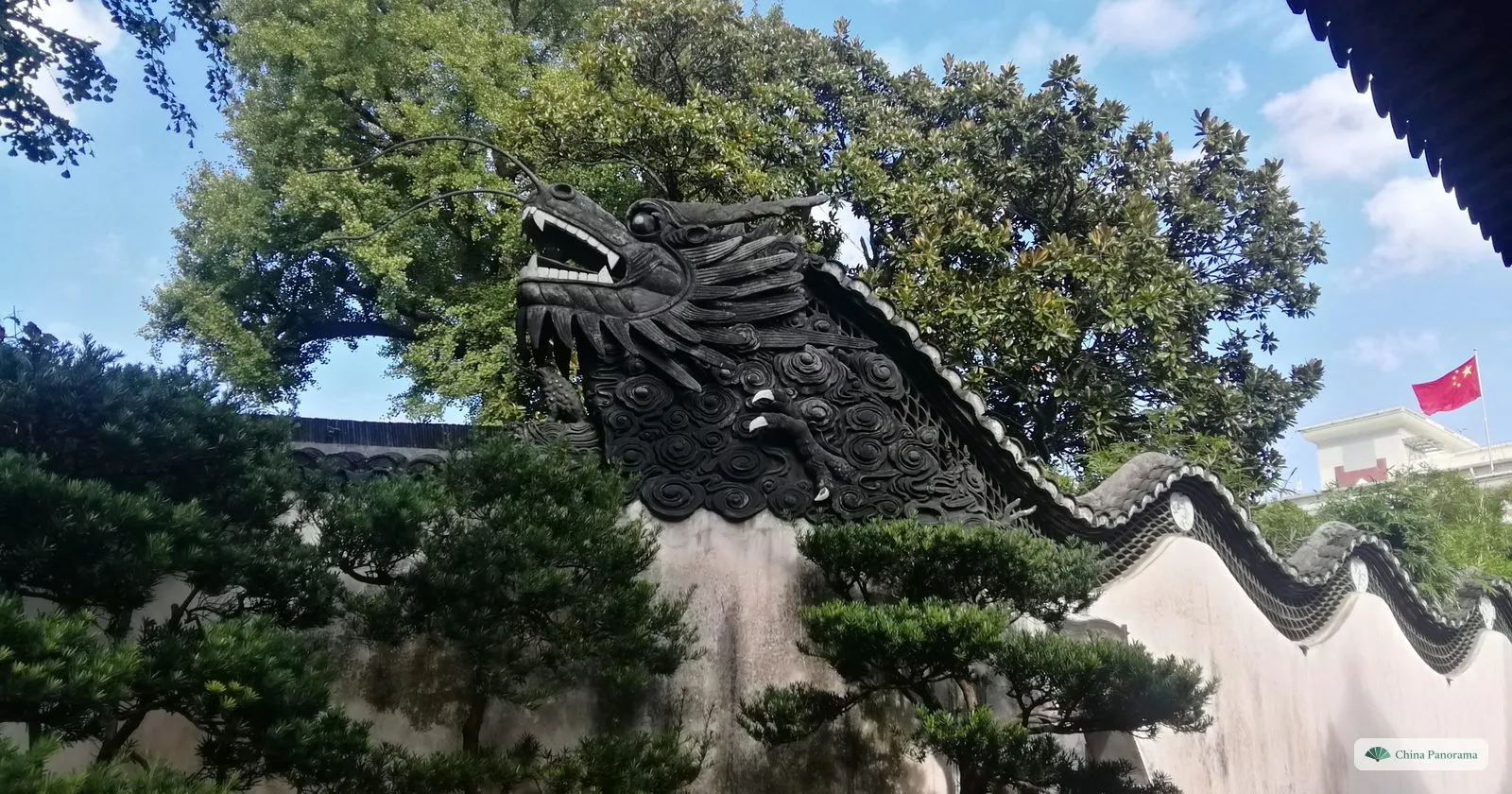 The head of a dragon carved at the end of a wavy whitewashed wall in Yu Garden, under trees and a blue sky