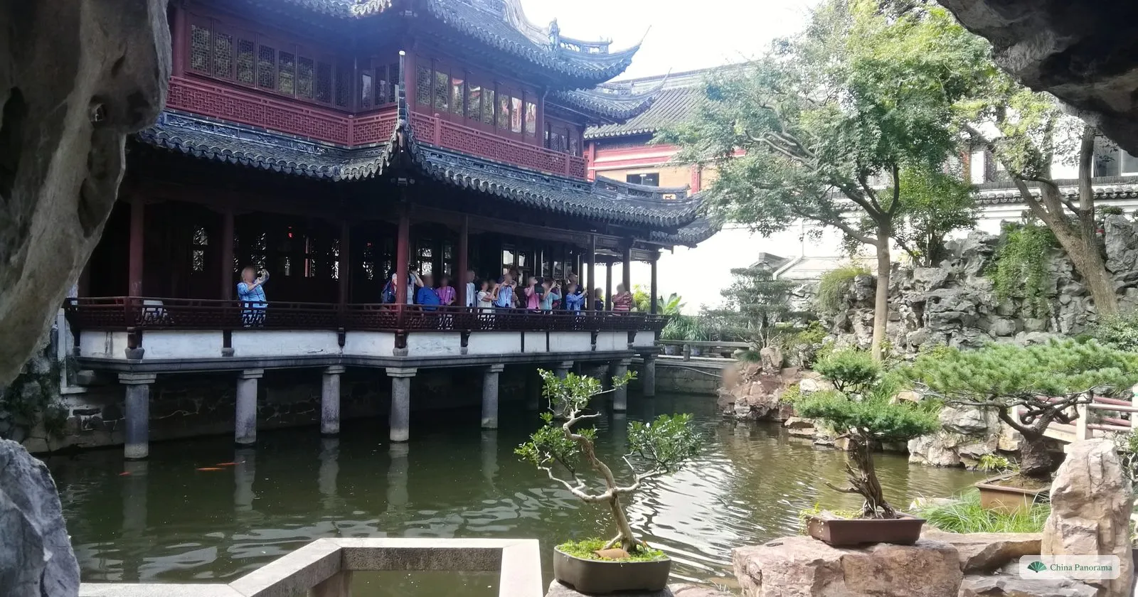 A two-storey hall on stilts over a pond in Yu Garden, with visitors on its balcony and bonsai trees on the rocks