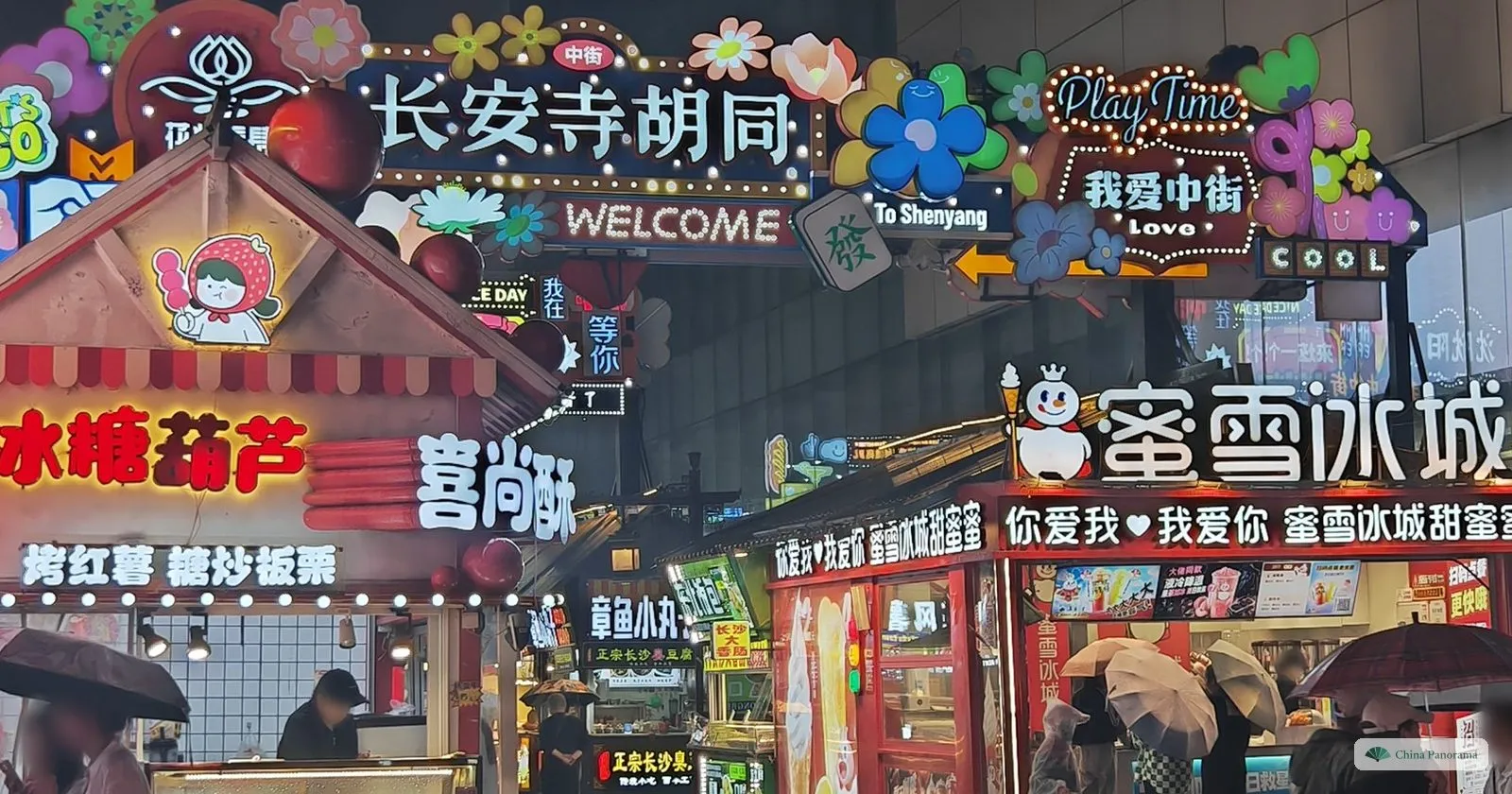 The neon entrance of Chang’ansi Hutong at night, a sign reading “Chang’ansi Hutong — WELCOME” above snack kiosks and people under umbrellas