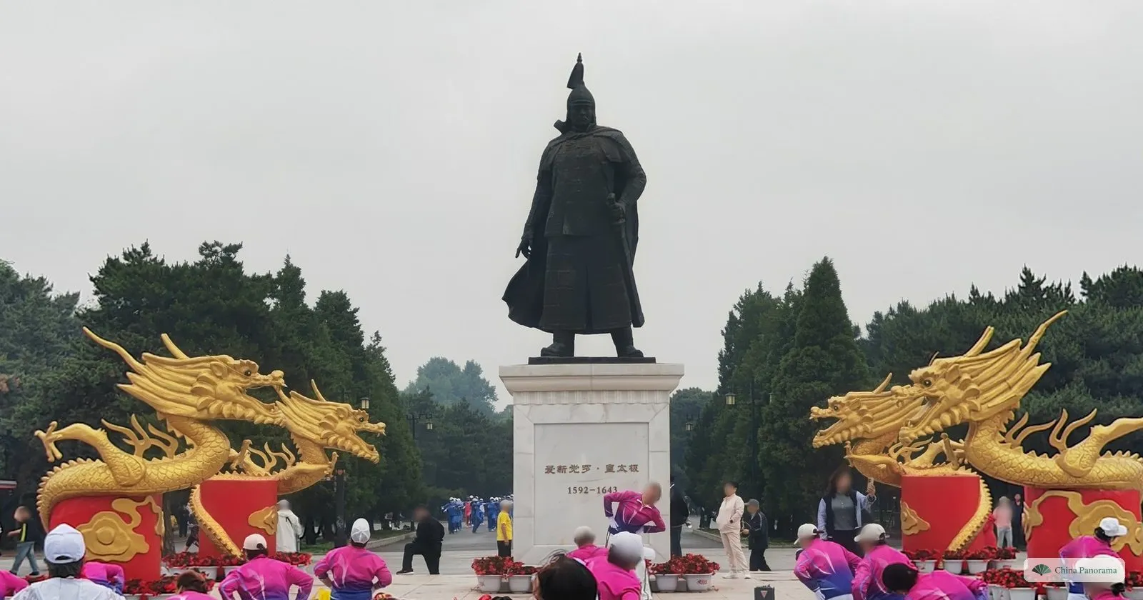 A statue of Hong Taiji in armour on a white plinth inscribed 1592–1643, between two golden dragons, with a dance group in pink tracksuits in front
