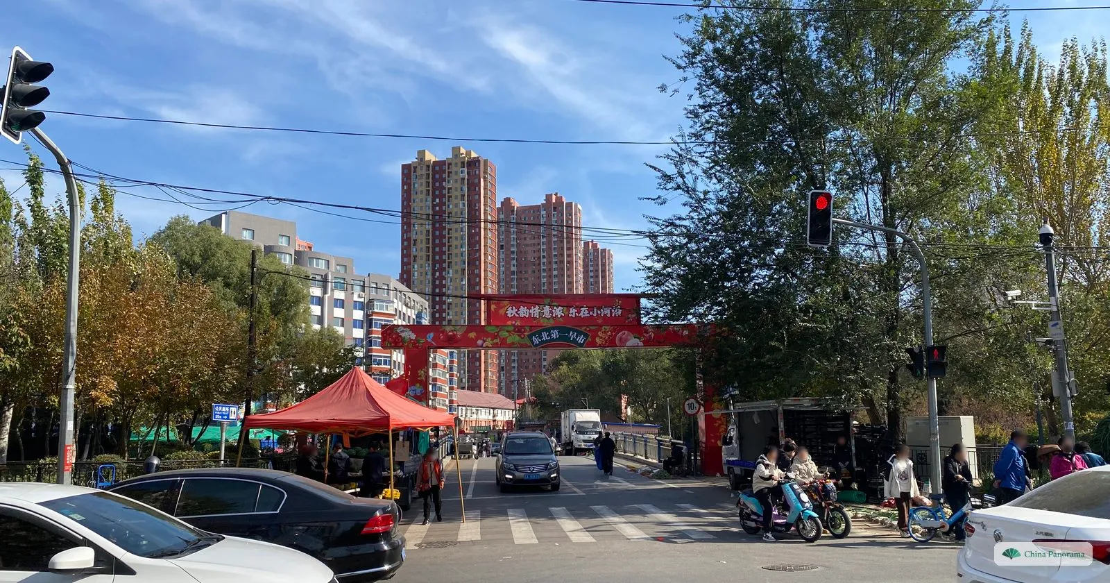 A red arch reading “No. 1 morning market of the Northeast” over a street in Shenyang, with a market tent, scooters and tower blocks under a blue sky