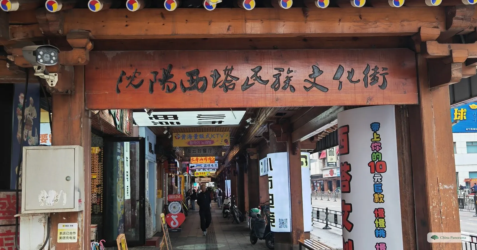 A wooden gateway with a carved sign reading “Shenyang Xita Ethnic Culture Street” over a covered pavement lined with shop signs
