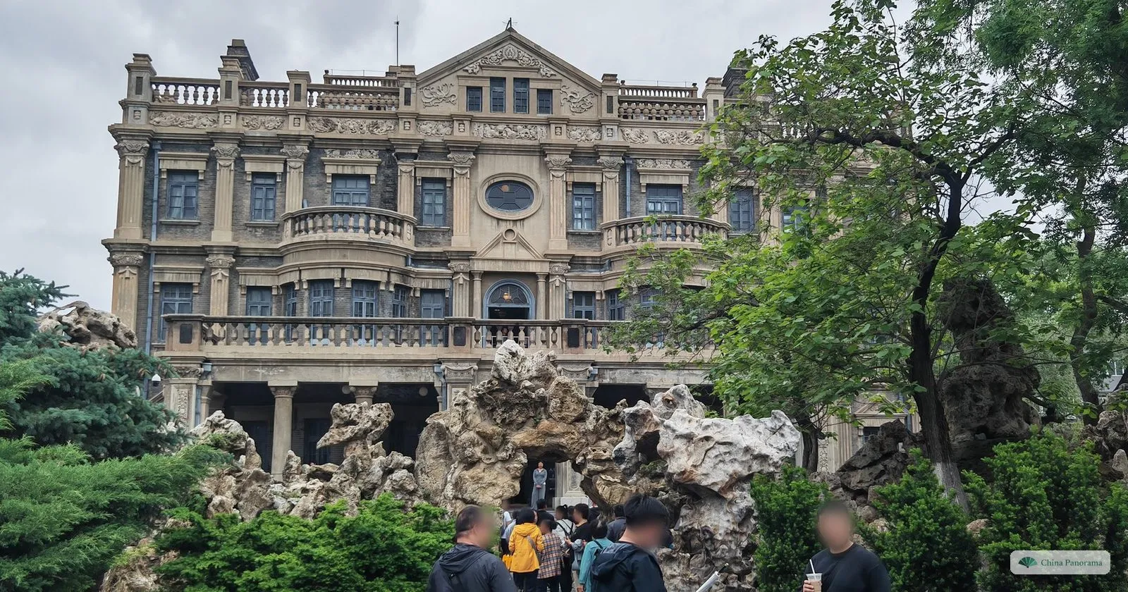 The grey stone Big Green Building at Zhang’s Mansion in Shenyang, behind a rockery, with visitors in front