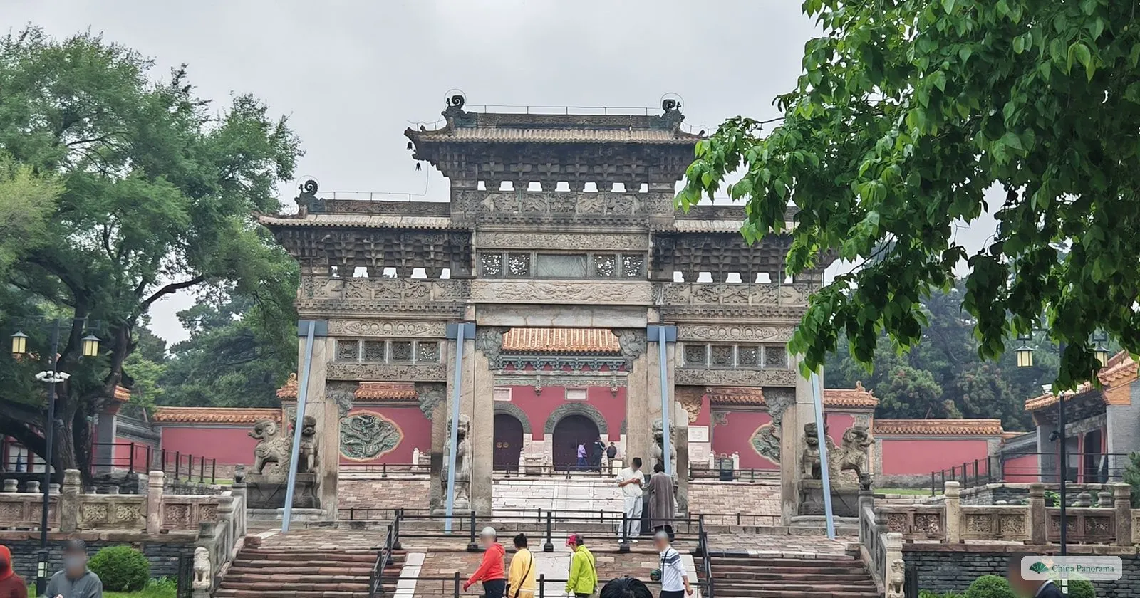 A carved grey stone archway with stone lions in front of the red walls of the Zhaoling tomb, propped with blue steel braces