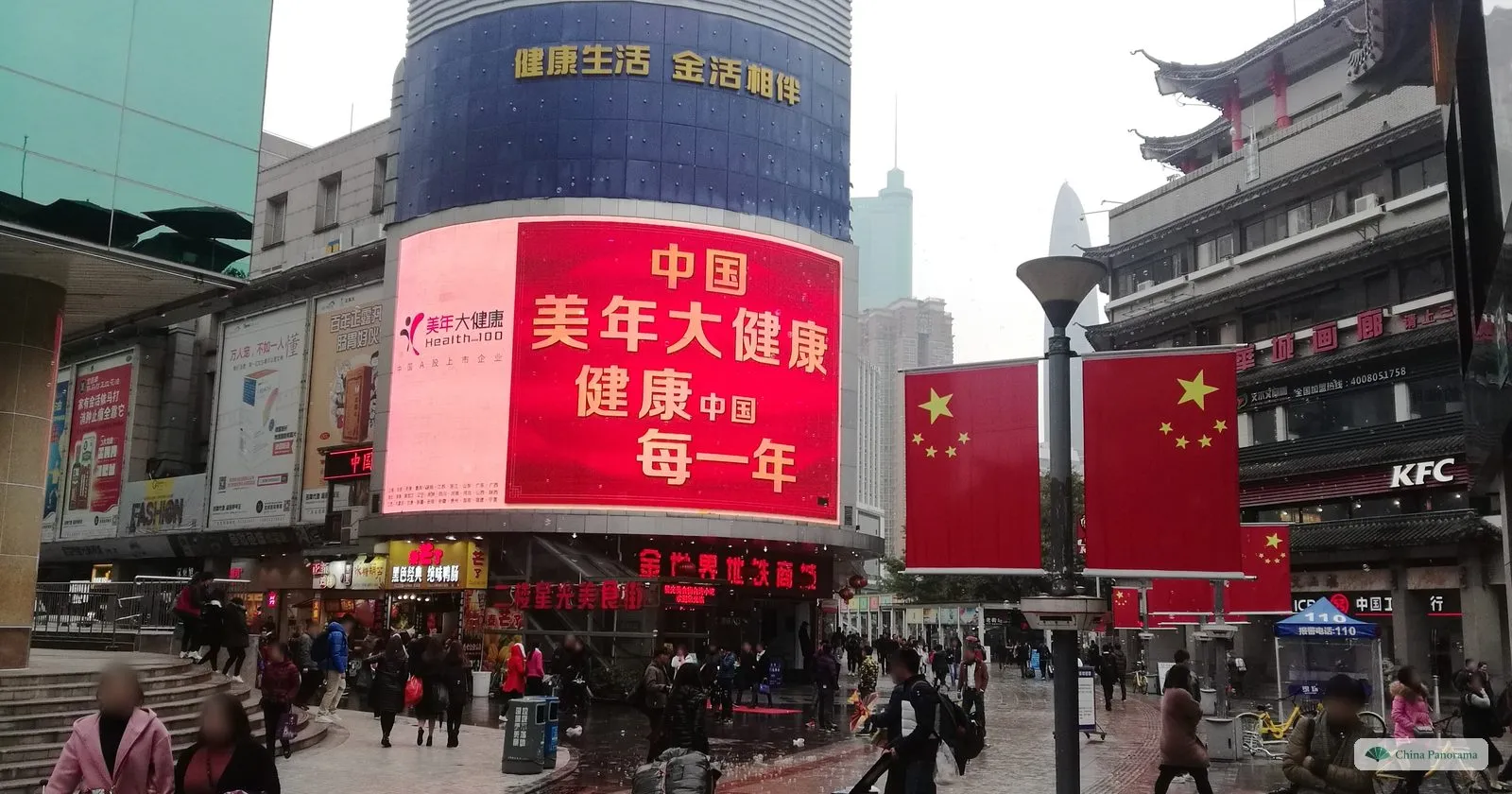 A busy corner in Dongmen with a huge curved LED screen showing red Chinese characters, Chinese flags on poles, a KFC and a crowd of shoppers