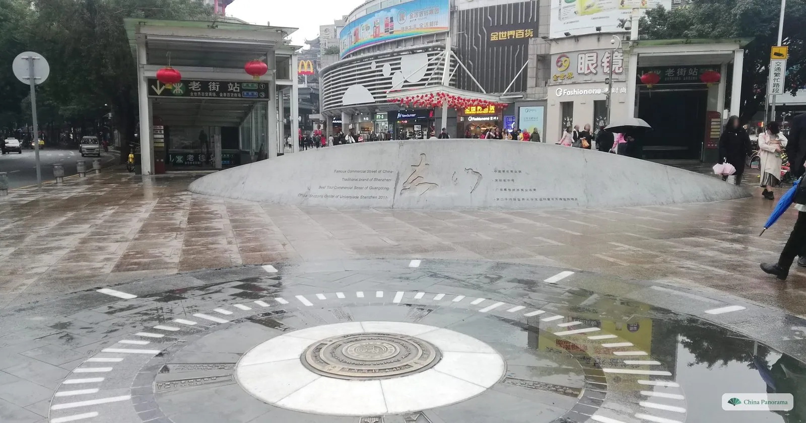 A round paved square in Dongmen with a large metal disc set in the wet ground, Chinese-style gateways and shops around it and people walking through