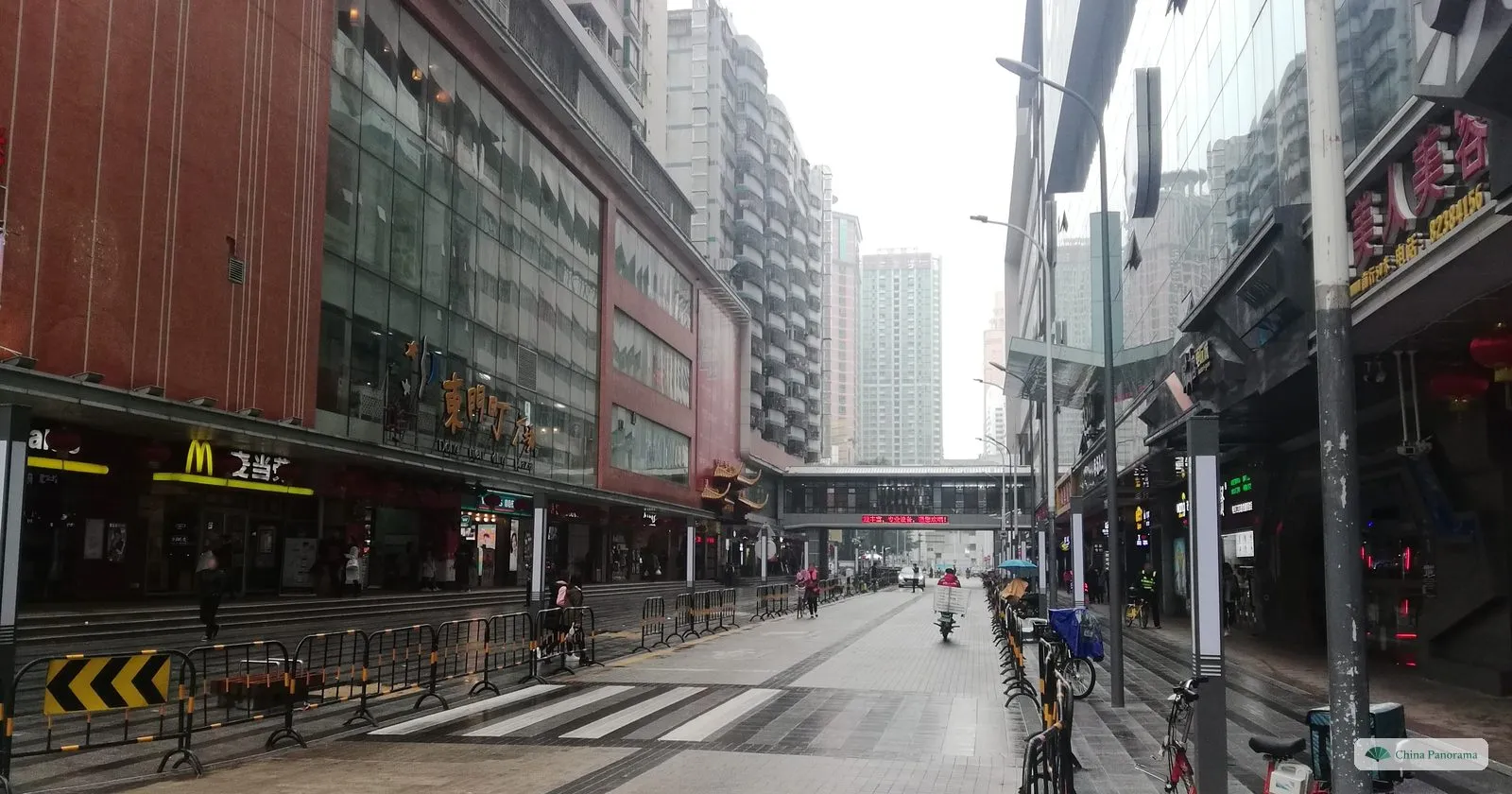 A wide pedestrian street in Dongmen on a wet day, with department stores on the left, a footbridge across the street and a few people with umbrellas