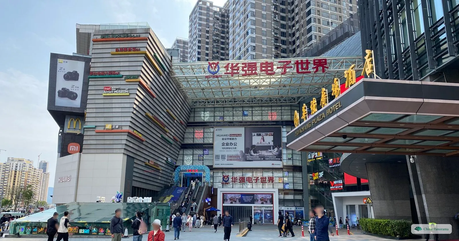 The entrance to Huaqiang Electronics World: a multi-storey mall with a red sign, escalators, shop fronts and people on the paved square in front