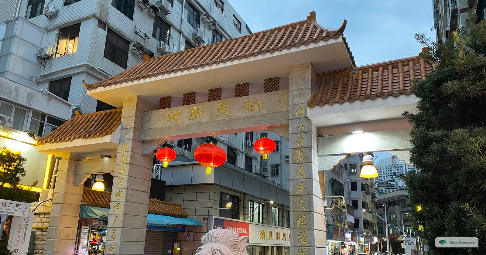 The granite gate of Hubei New Village with orange tiled roofs, red lanterns and the gilded name 湖贝新村, between apartment blocks at dusk