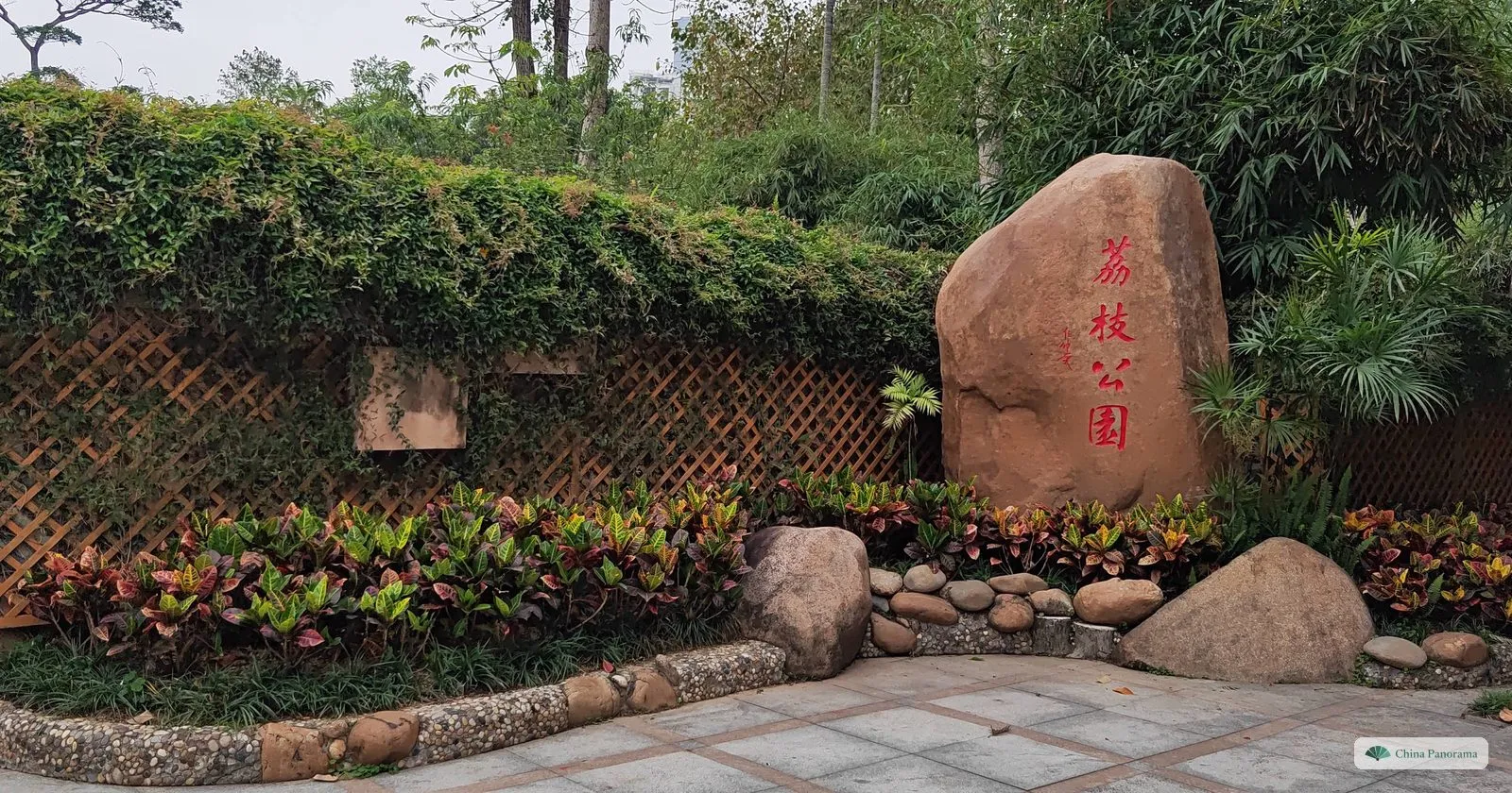 A tall red boulder carved with the characters 荔枝公园 at the entrance to Lizhi Park, set among flower beds, rocks and a wall covered in creepers