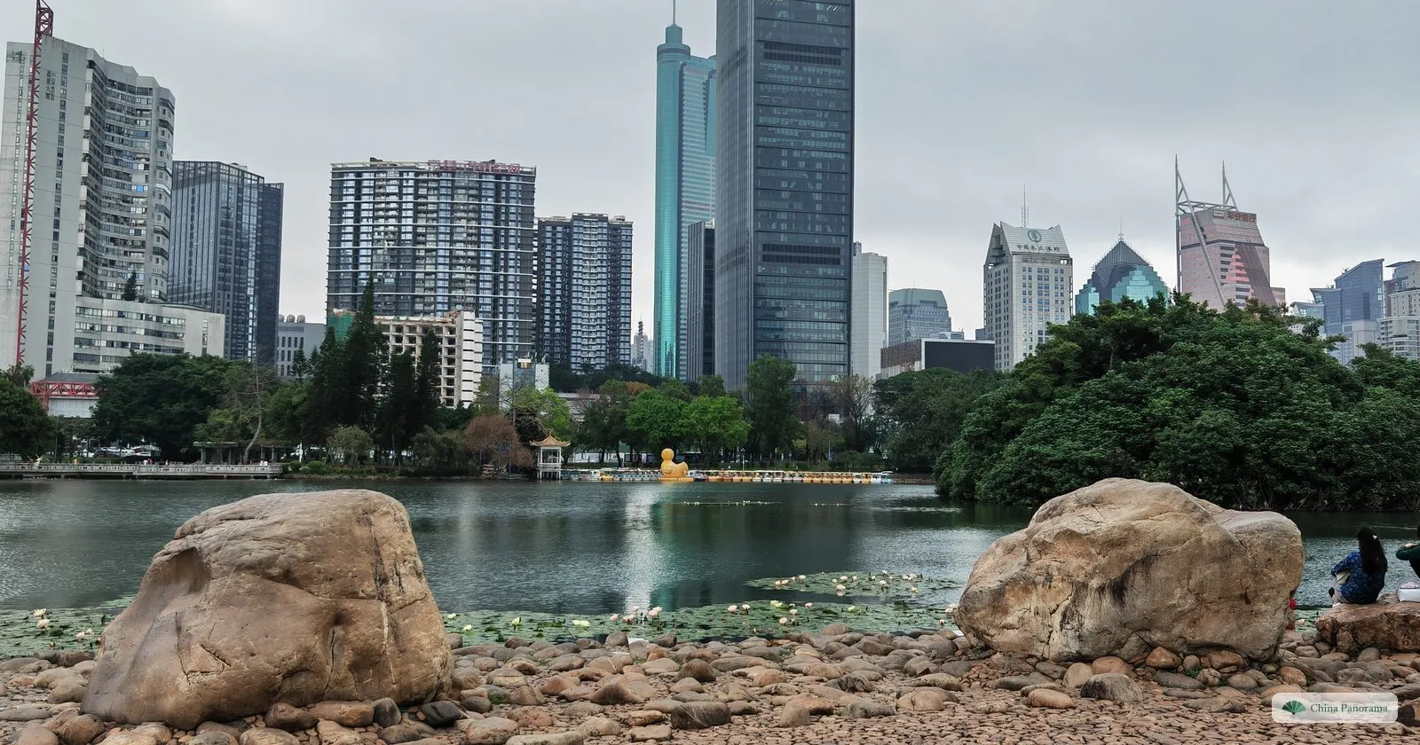 Rounded boulders on the shore of the lake in Lizhi Park, a wooded island in the water, and a row of high-rise towers with spires behind it under grey cloud