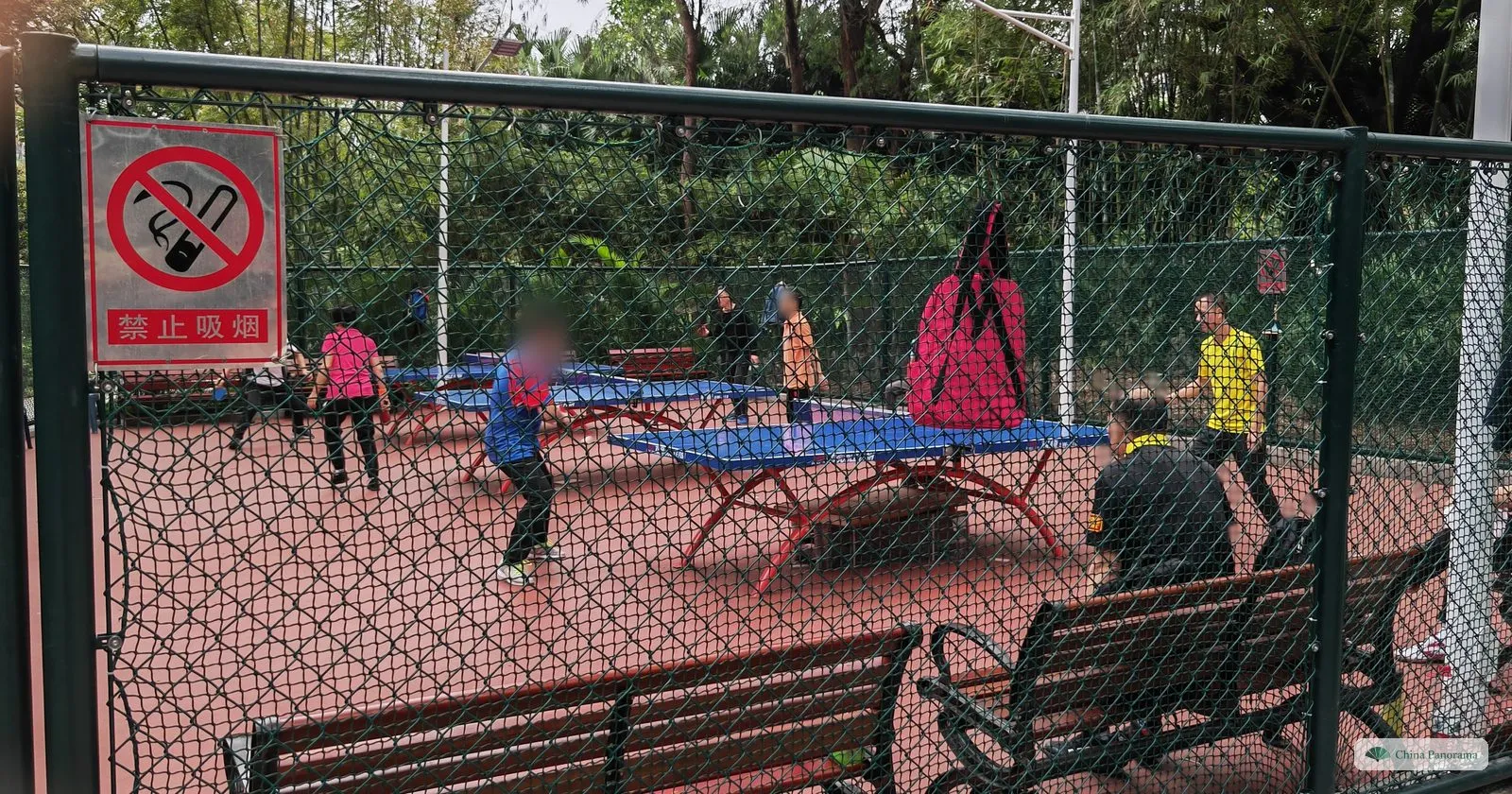 Outdoor table tennis tables on a red court behind a wire fence in Lizhi Park, a no-smoking sign on the fence and several people playing