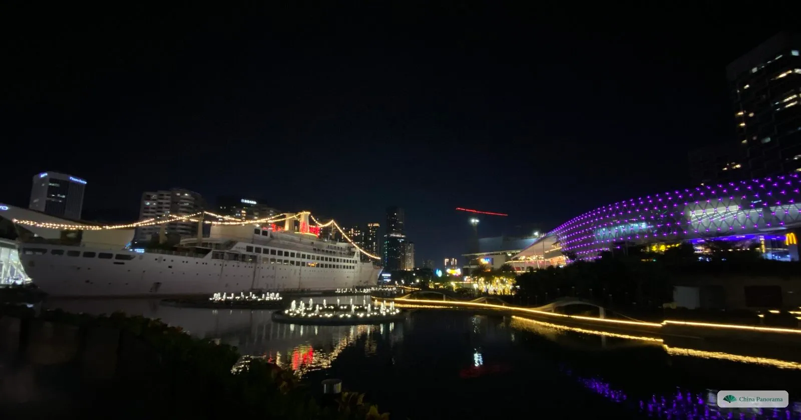 The Minghua at night across dark water, its decks outlined with lights, with a purple-lit building and apartment towers to the right
