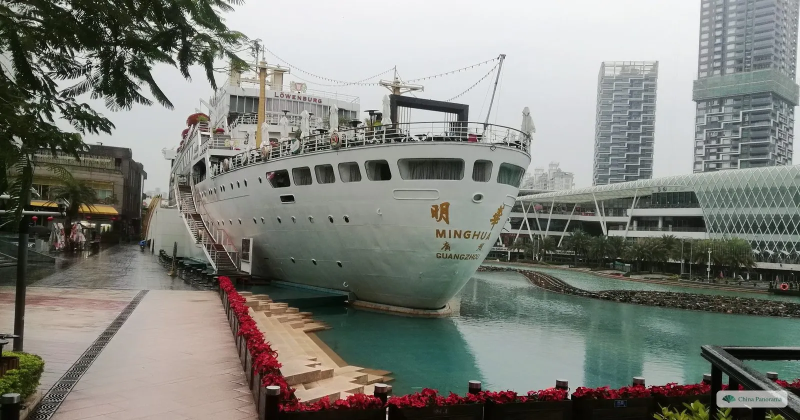 The white cruise liner Minghua moored in a turquoise basin at Sea World on a rainy morning, its name in Chinese and Latin letters on the bow, a wet promenade beside it