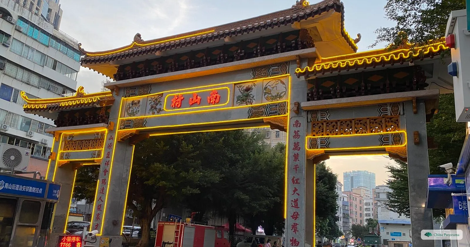 The gate of Nanshan Village at dusk: a three-bay archway with glazed roofs outlined in yellow lights, the name 南山村 in red characters and couplets on the pillars