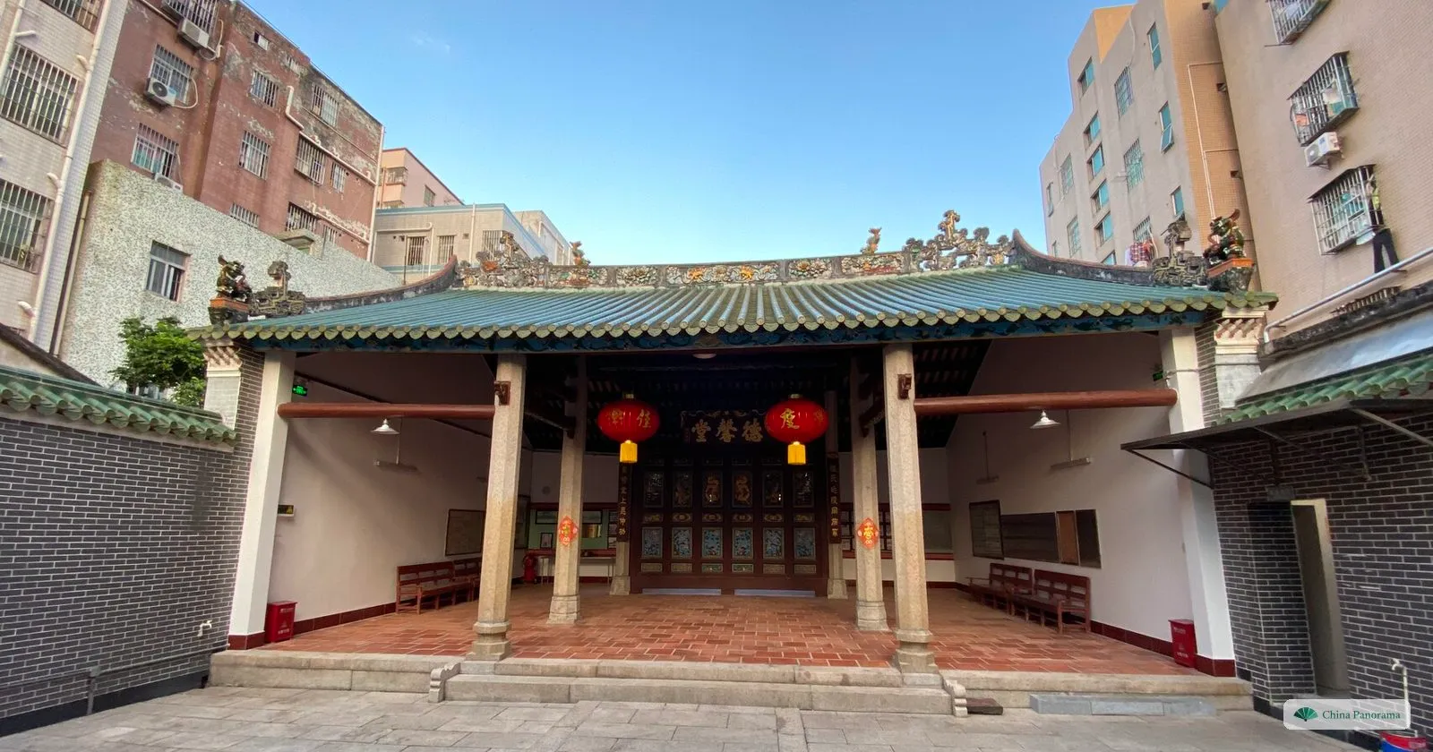 The courtyard of Dexin Hall under a blue sky, its green-tiled hall with red lanterns and benches along the walls, between apartment blocks