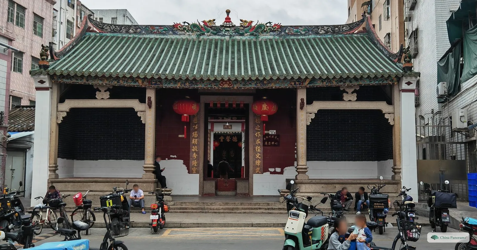 The front of Dexin Hall in Nanyuan village: a green-tiled roof with dragons on the ridge, granite columns, dark brick walls, red lanterns and gilded couplets, with men seated on the steps and scooters parked in front