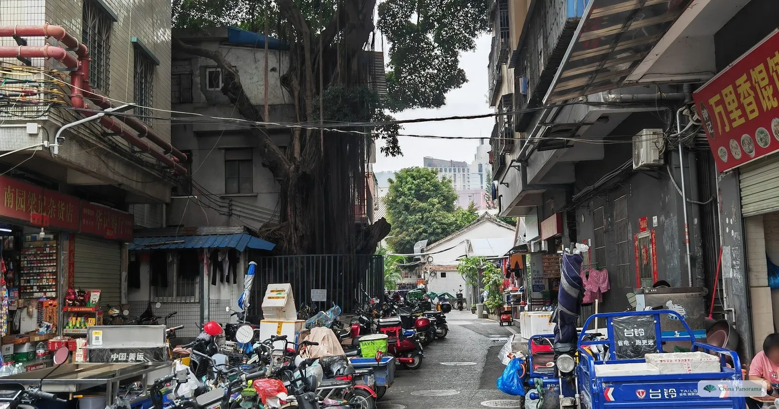 A cluttered lane in Nanyuan village with a shop sign, piles of goods, parked scooters and a banyan tree behind the buildings
