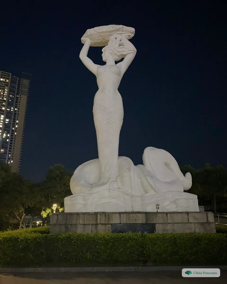 The white statue of the goddess Nüwa at night, holding a stone above her head, her body ending in a coiled serpent tail on a wave-shaped base
