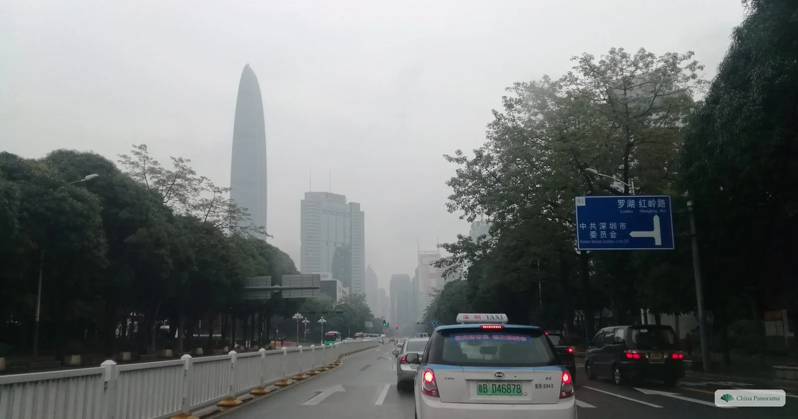 The tapering Ping An Finance Centre in the distance on a misty day, its top fading into cloud, seen down a wide road lined with trees, a taxi in front